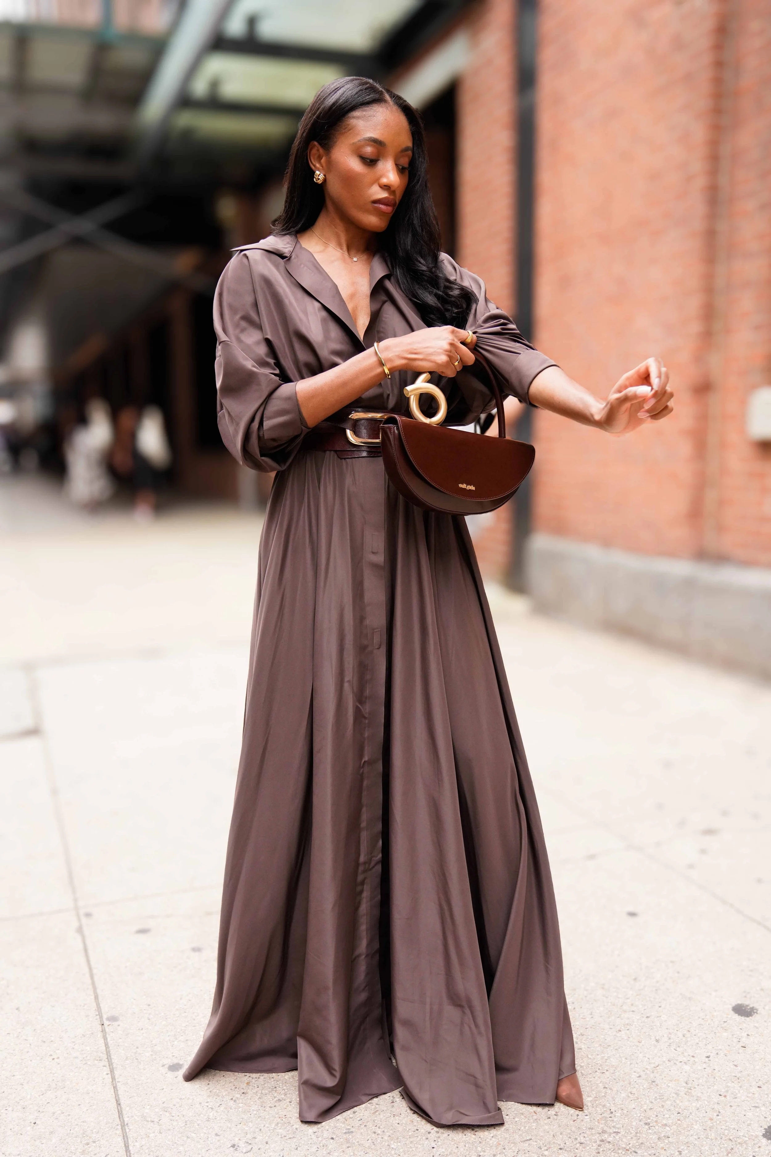 Fashionable woman in a long brown dress with a matching belt, carrying a small dark brown handbag, standing on a city sidewalk near brick buildings.