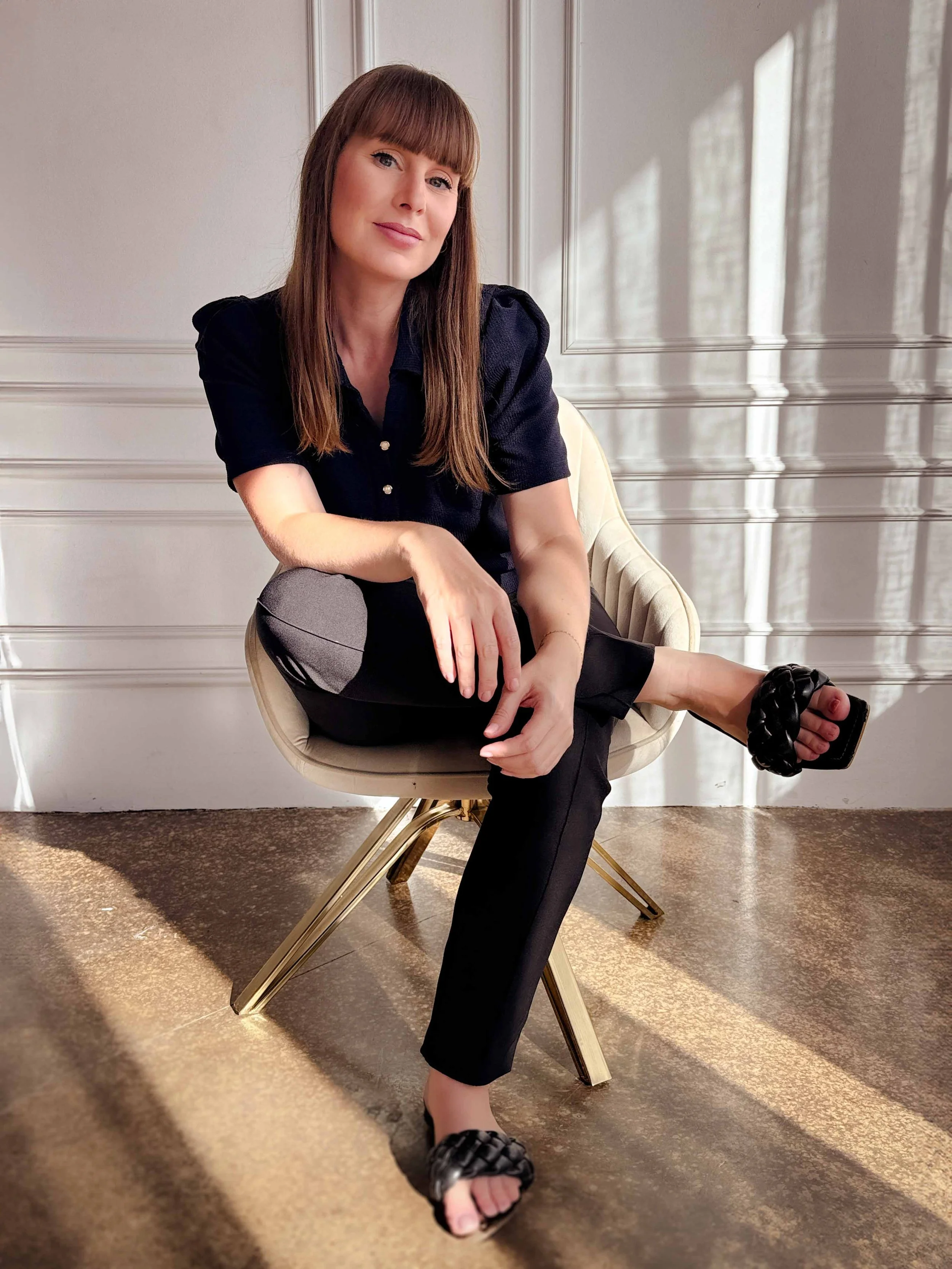 A woman with long brown hair and bangs sitting on a cream-colored chair near a wall with sunlight casting shadows. She is wearing a black blouse with short puffed sleeves, black pants with gray patches, black braided sandals, and has a gentle smile.