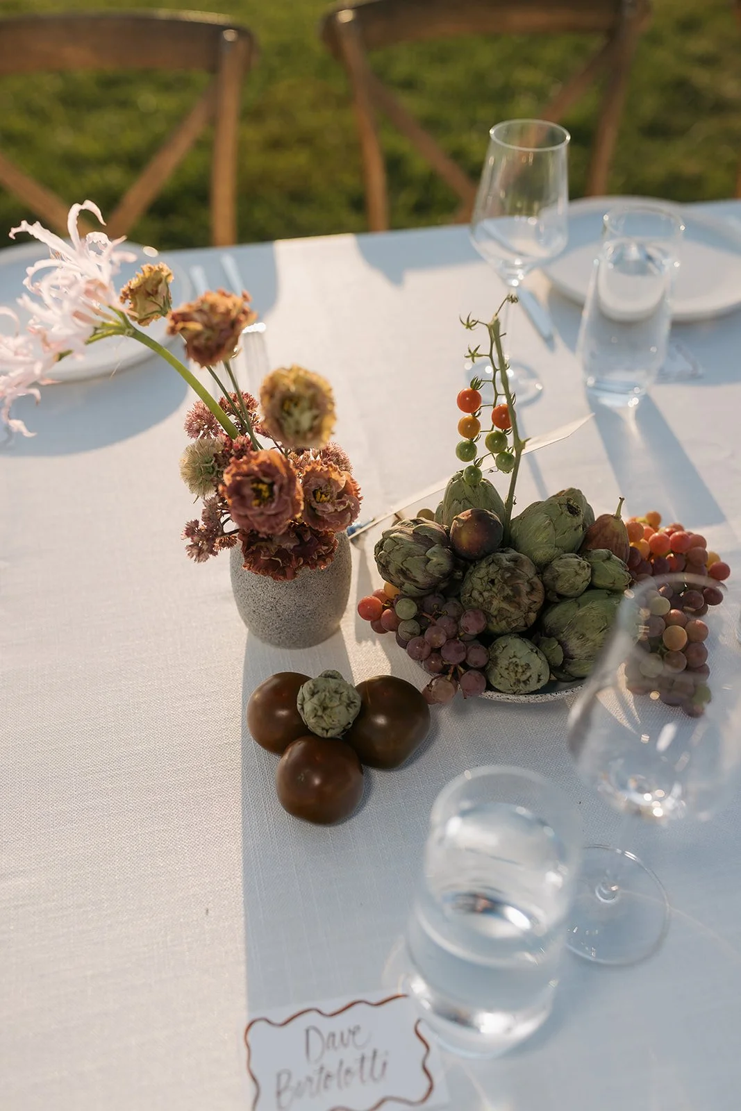 A white tablecloth decorated with a small vase of pink and brown flowers, a bowl of artichokes and grapes, and a cluster of chocolates, set outdoors on a sunny day with glassware and a handwritten name tag.