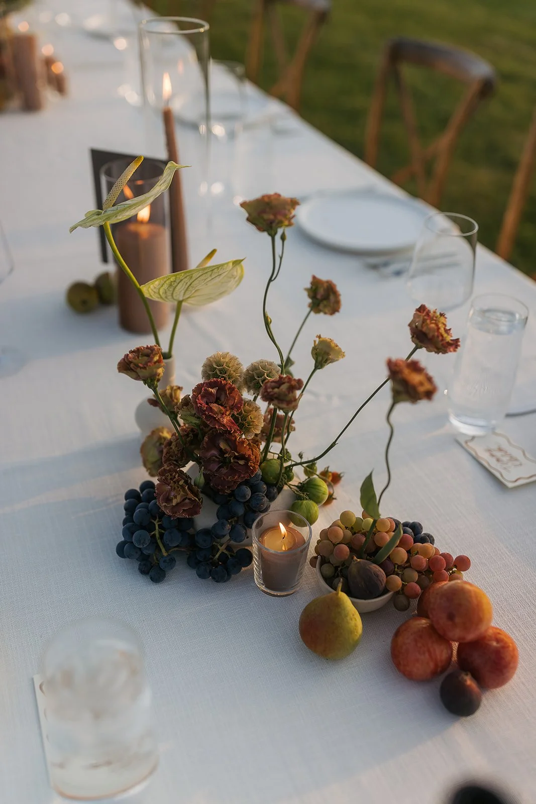 A decorated dining table with floral arrangements, grapes, pears, pomegranates, candles, and glassware at an outdoor event during sunset.