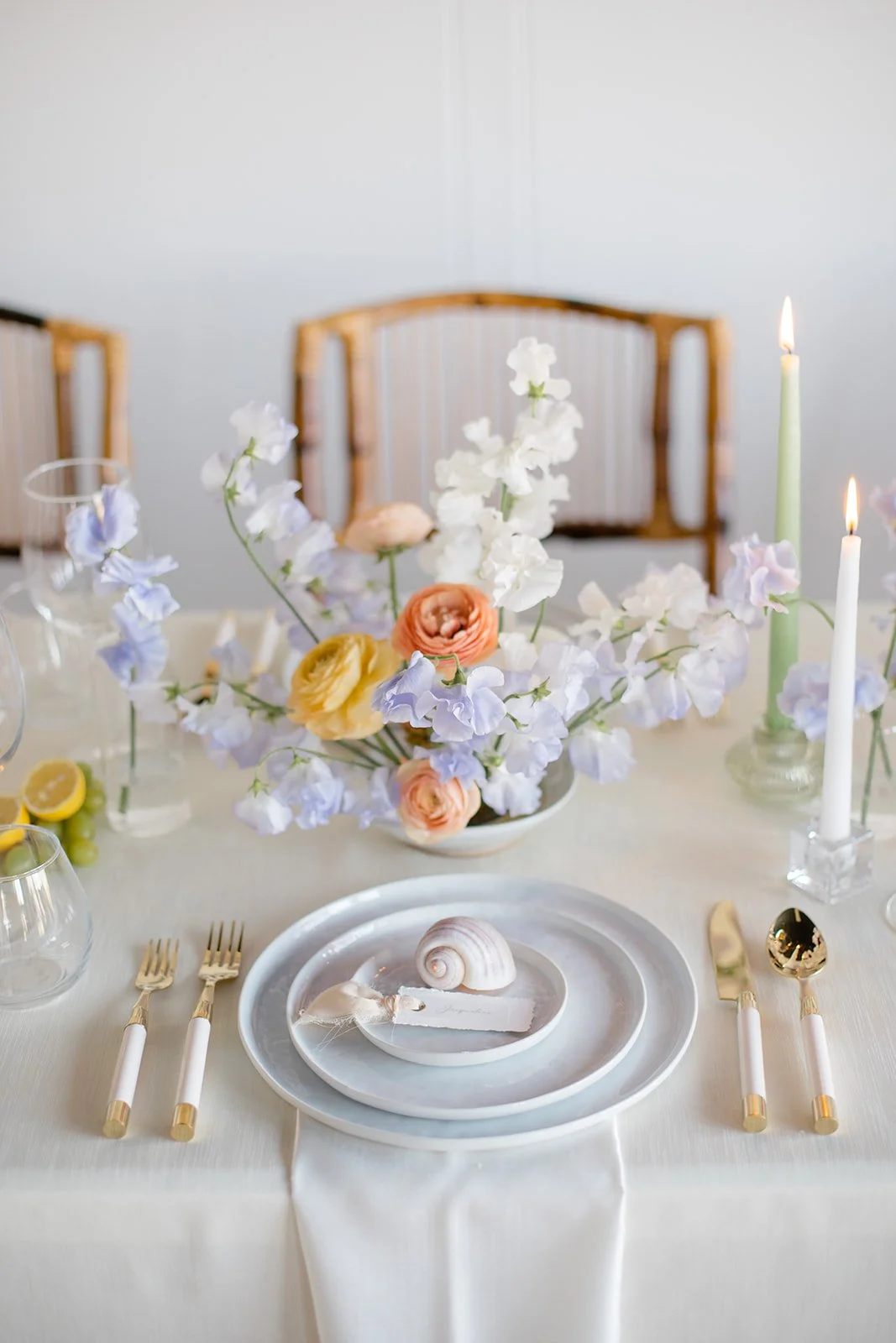 A table setting with white plates, gold and white cutlery, a seashell decoration, and a floral centerpiece with white, purple, yellow, and peach flowers. There are two lit candles and a chair with a wooden back in the background.