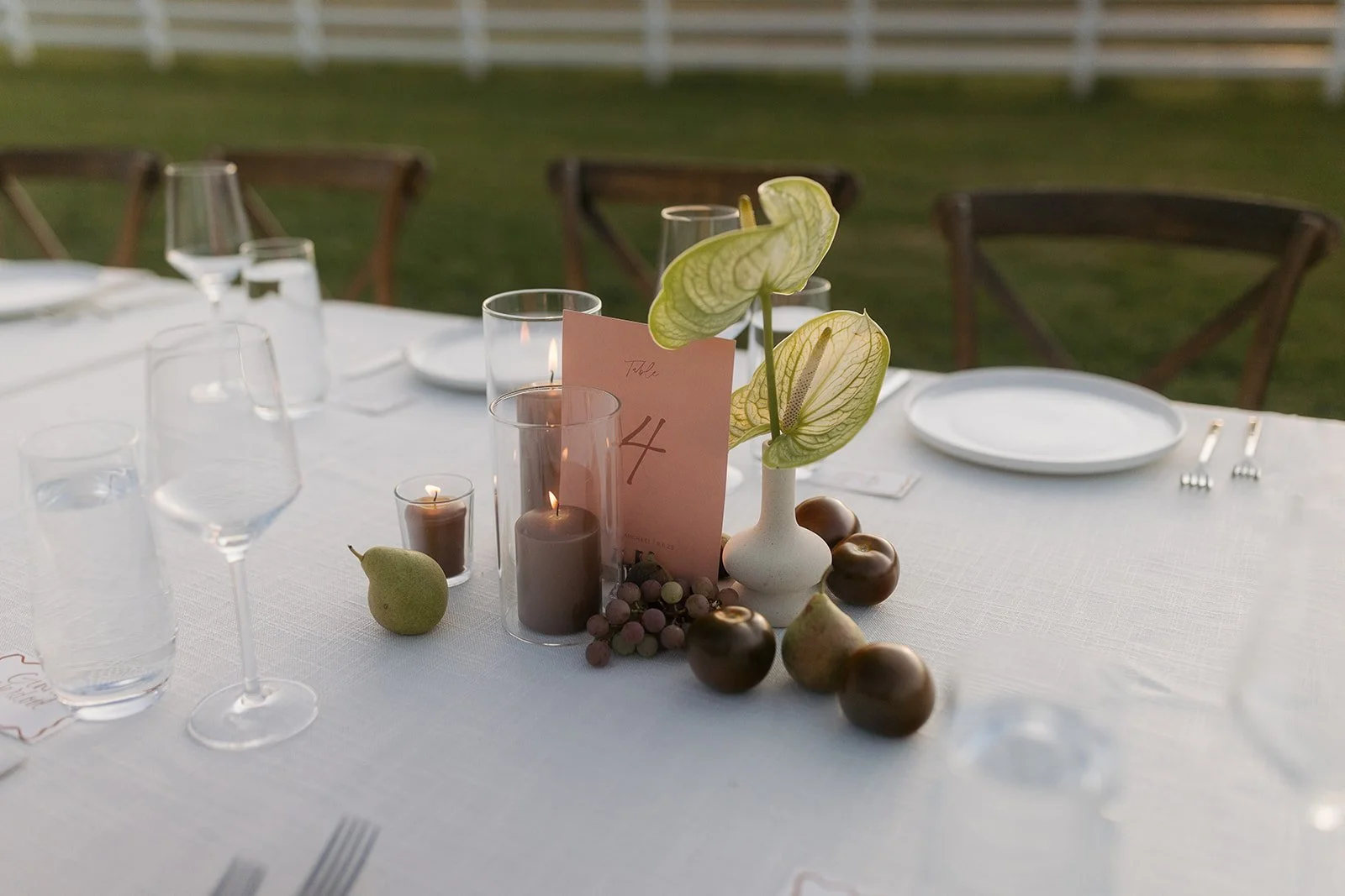 An outdoor dining table decorated with candles, pears, figs, a potted plant, and a table number card.