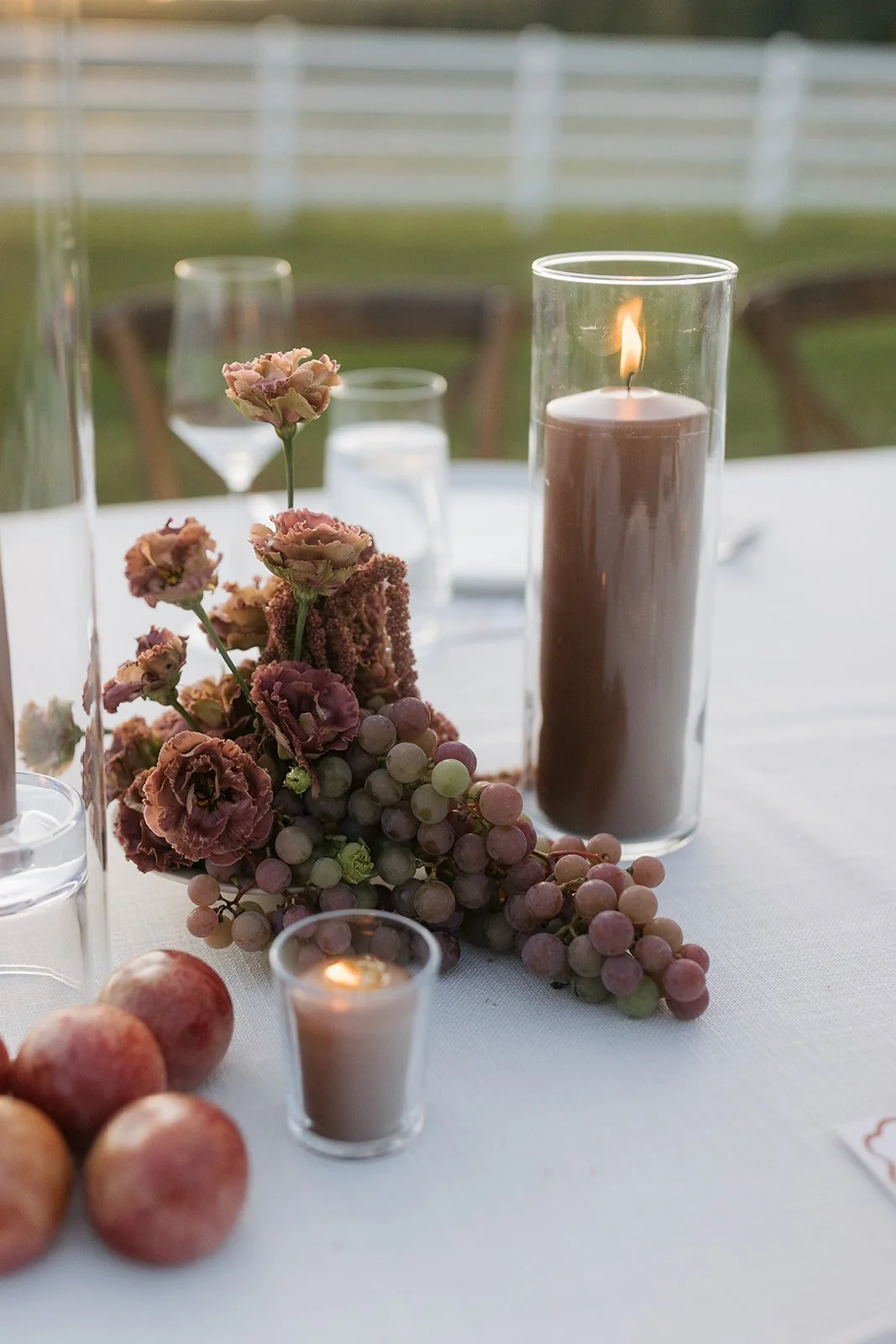 A table setting with candles, a floral arrangement, grapes, and peaches outdoors.