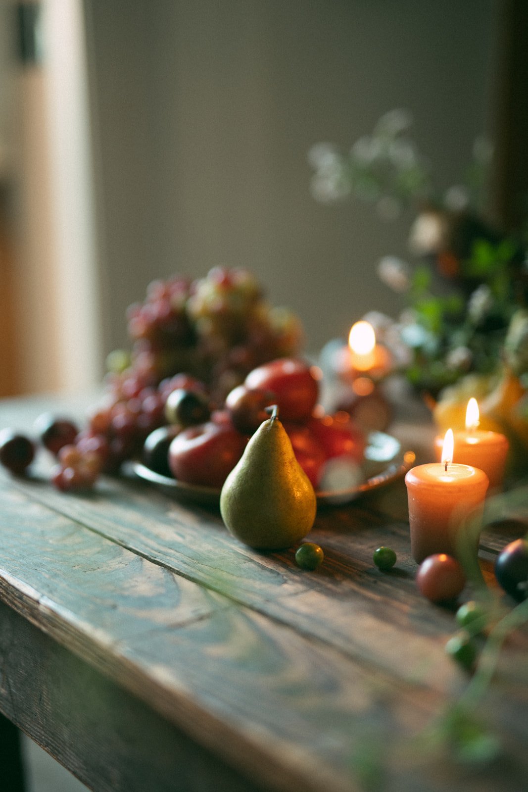 A still life of fruits including grapes, apples, a pear, and some small round fruits on a rustic wooden table, illuminated by candles, with a blurred background.