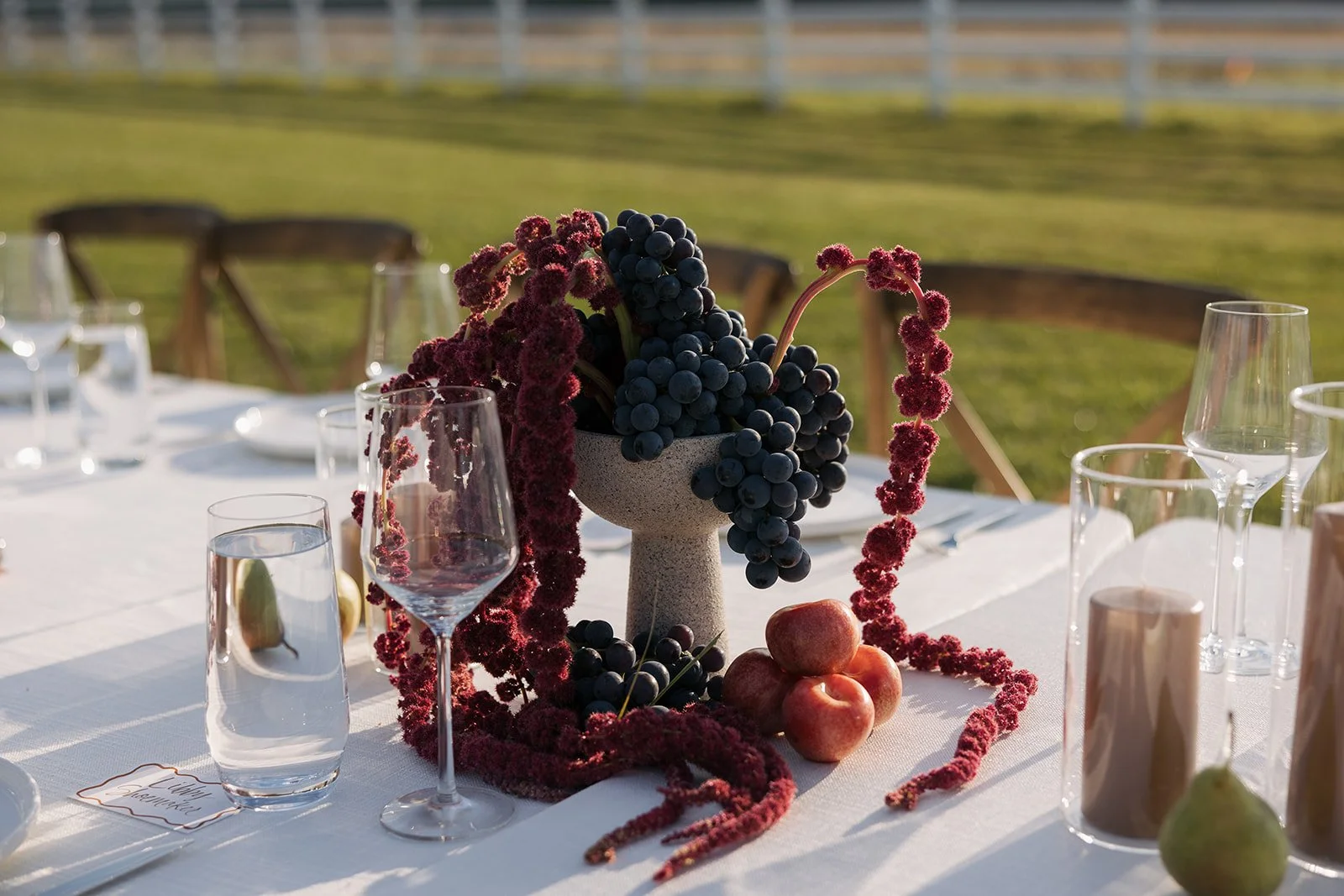 Table decorated with a centerpiece of grapes, red berries, and apples, with glasses, candles, and place settings outdoors.