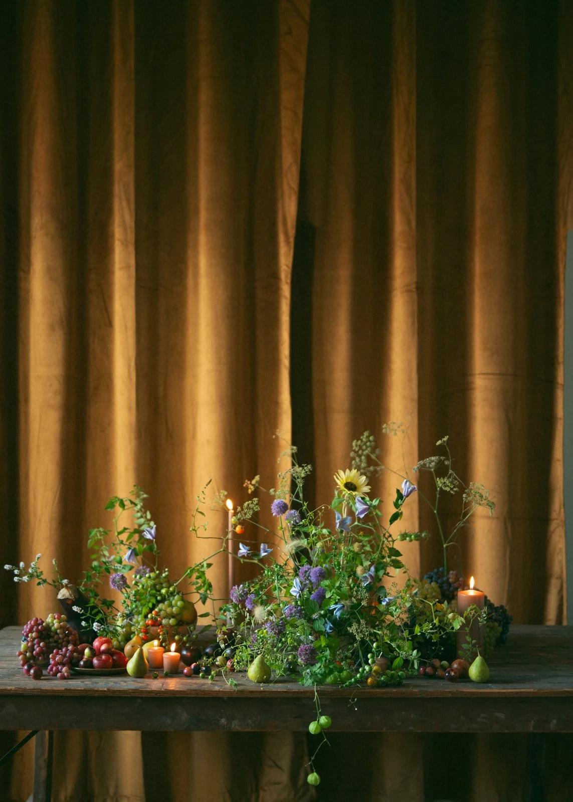 A wooden table with a large floral arrangement, various fruits, and small lit candles in front of a gold curtain.