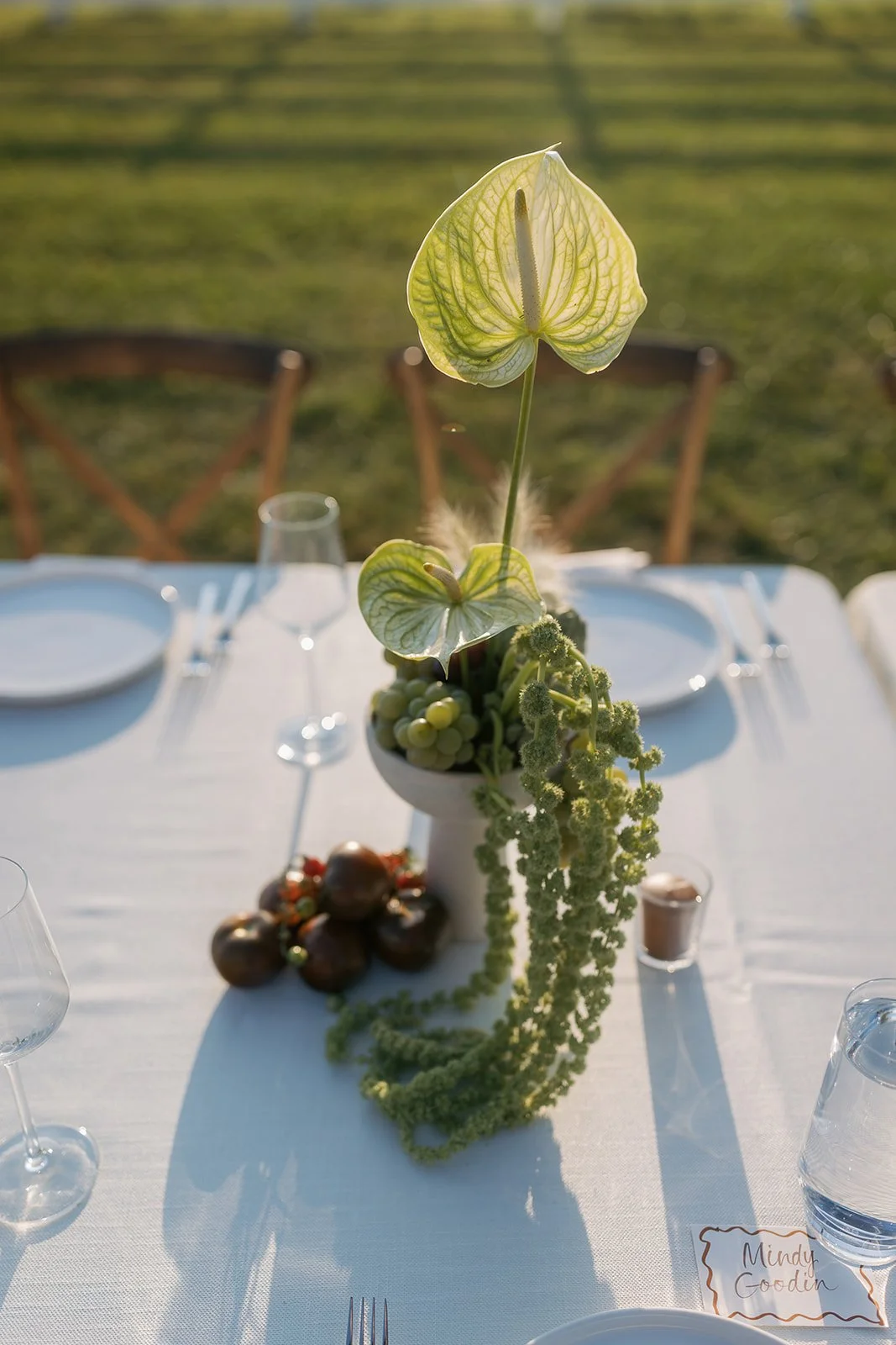 A tall floral arrangement with large green and white anthurium flowers and trailing green foliage on a white tablecloth at an outdoor event, with chairs, glasses, and plates in the background.