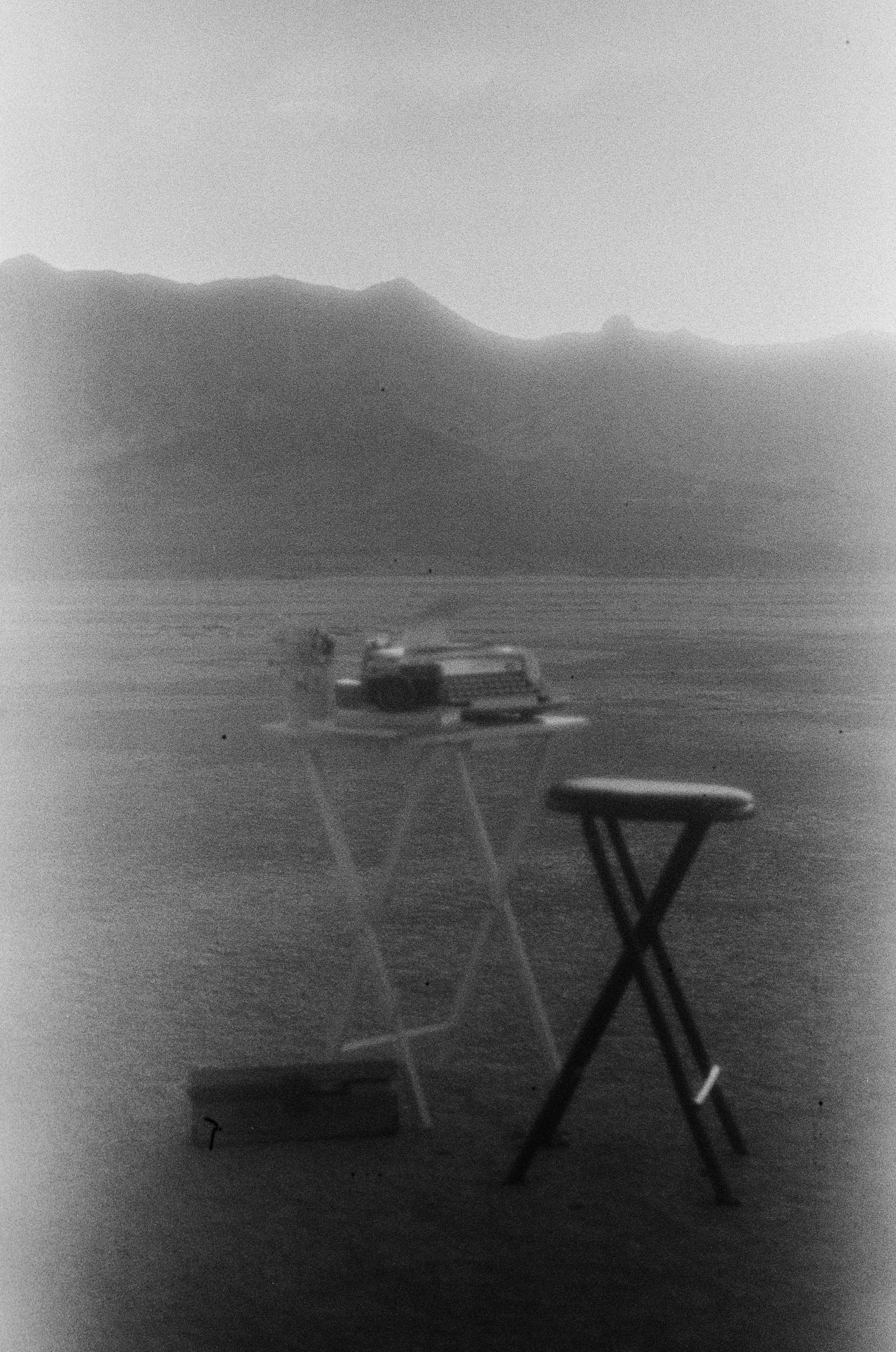 A black-and-white photograph of a beach scene with a mountain range in the background. In the foreground, there is a foldable stool, a small table with an object on it, and some other indistinct items.
