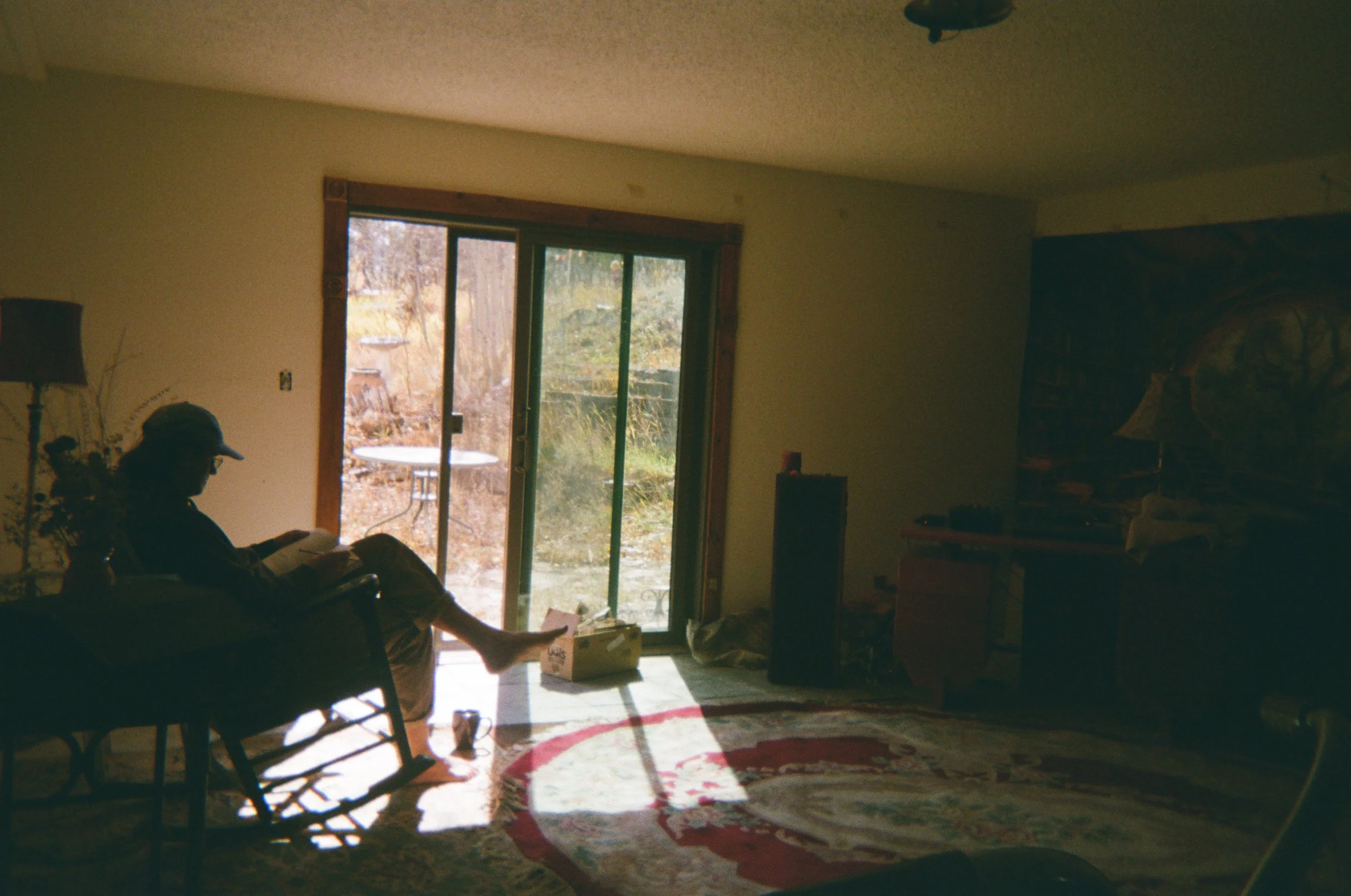 Person sitting on a couch reading a book in living room with sunlight coming through glass sliding door to outdoor patio.