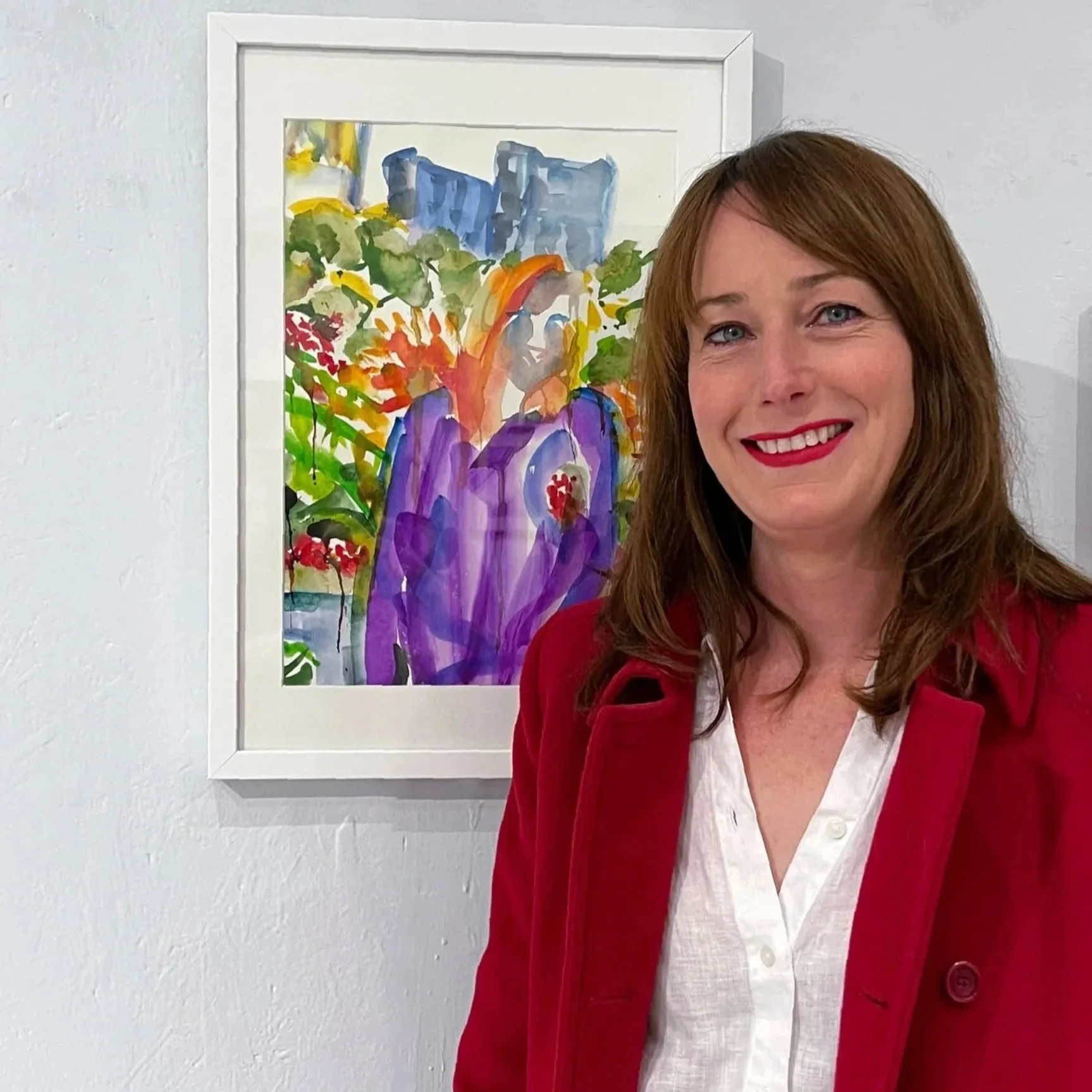 A woman with shoulder-length auburn hair and red lipstick, smiling, stands in front of a white wall with a framed colorful watercolor painting of a person surrounded by flowers and greenery.