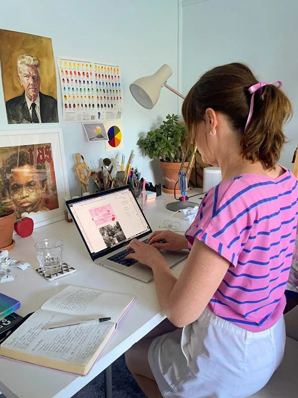 A woman with brown hair tied back with a pink ribbon, wearing a pink and purple striped shirt and white shorts, sitting at a desk working on a laptop in a brightly decorated art studio or creative workspace.