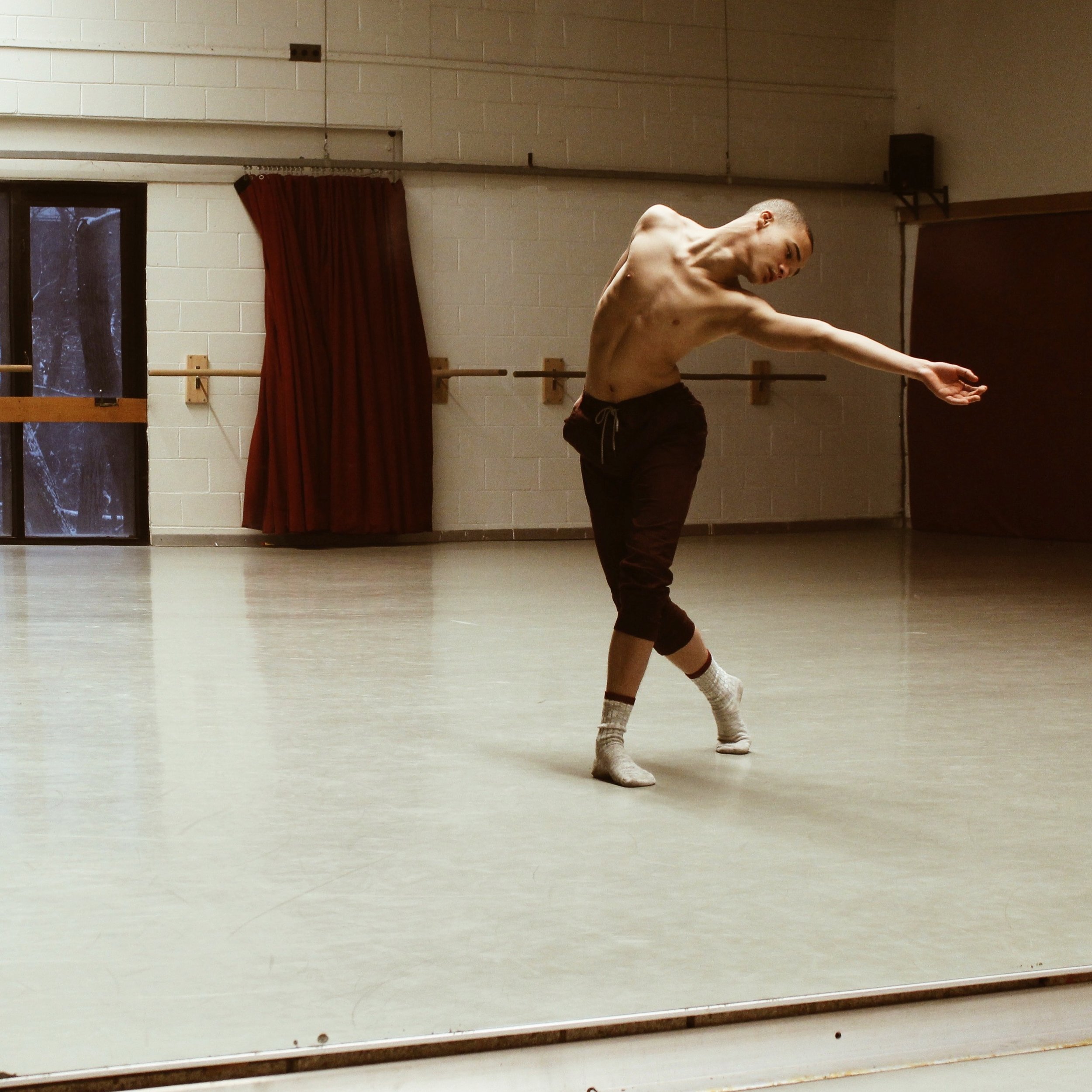 A shirtless man dancing alone in a dance studio with a mirror, red curtains, and ballet barres.