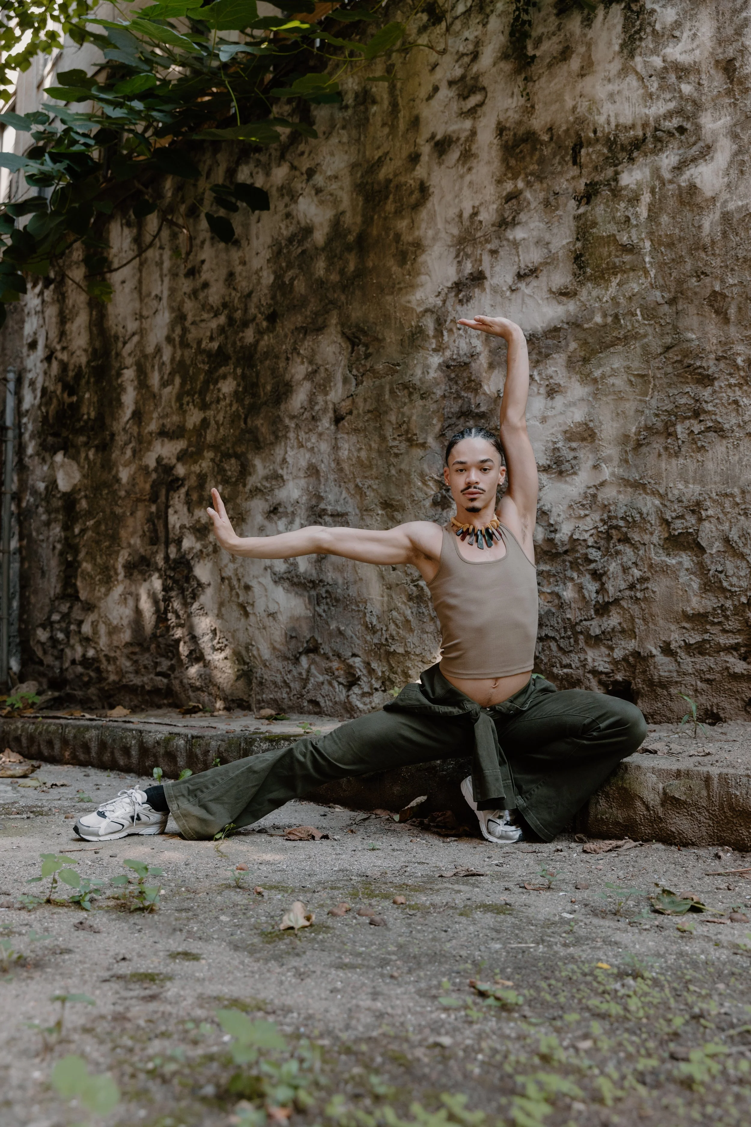 A person with long hair tied back in a bun practicing yoga outdoors against a weathered wall, performing a side lunge with arms extended in a graceful pose.