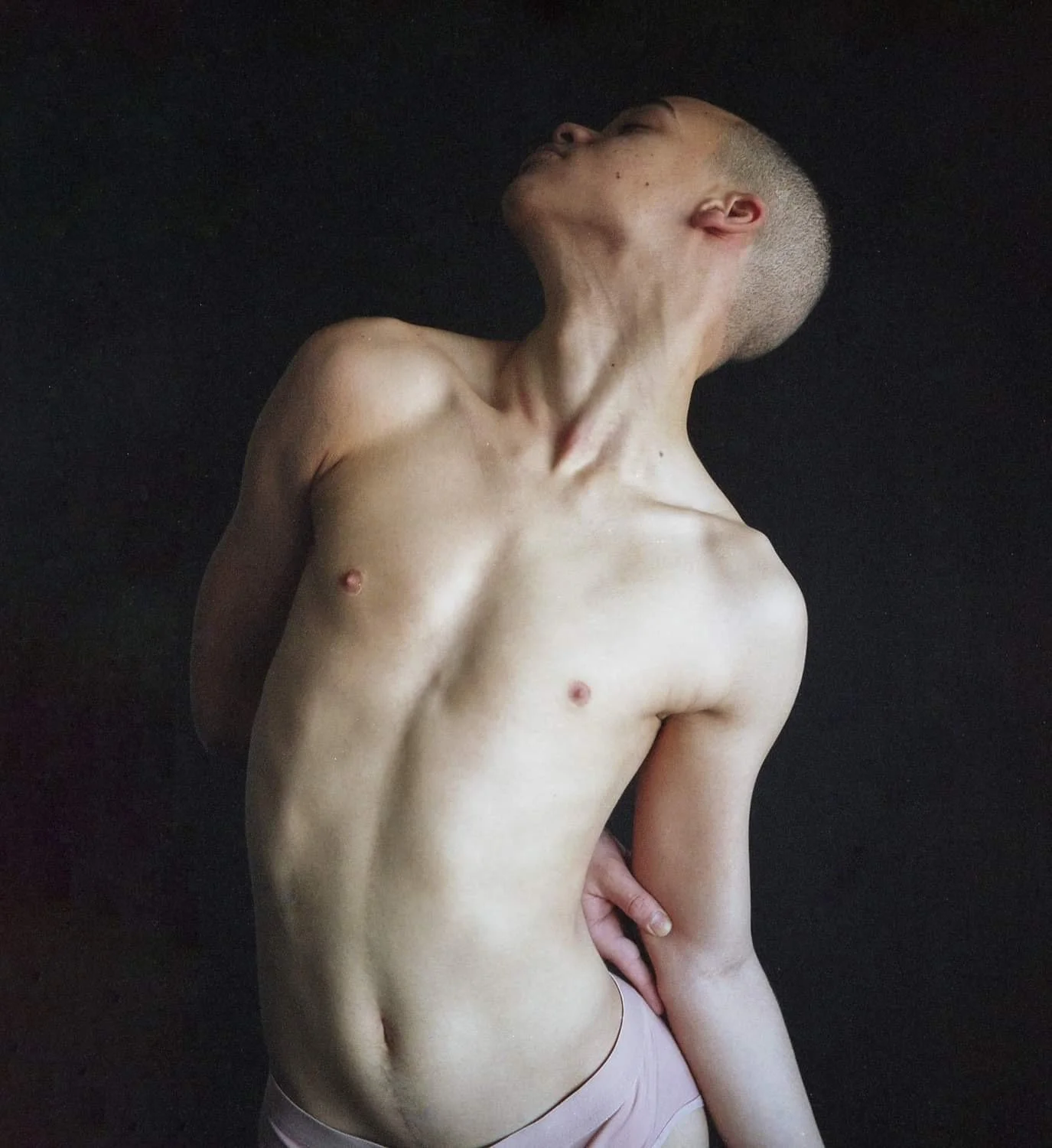 A nude person with a shaved head, posing against a black background, arching their neck back and tilting their head.