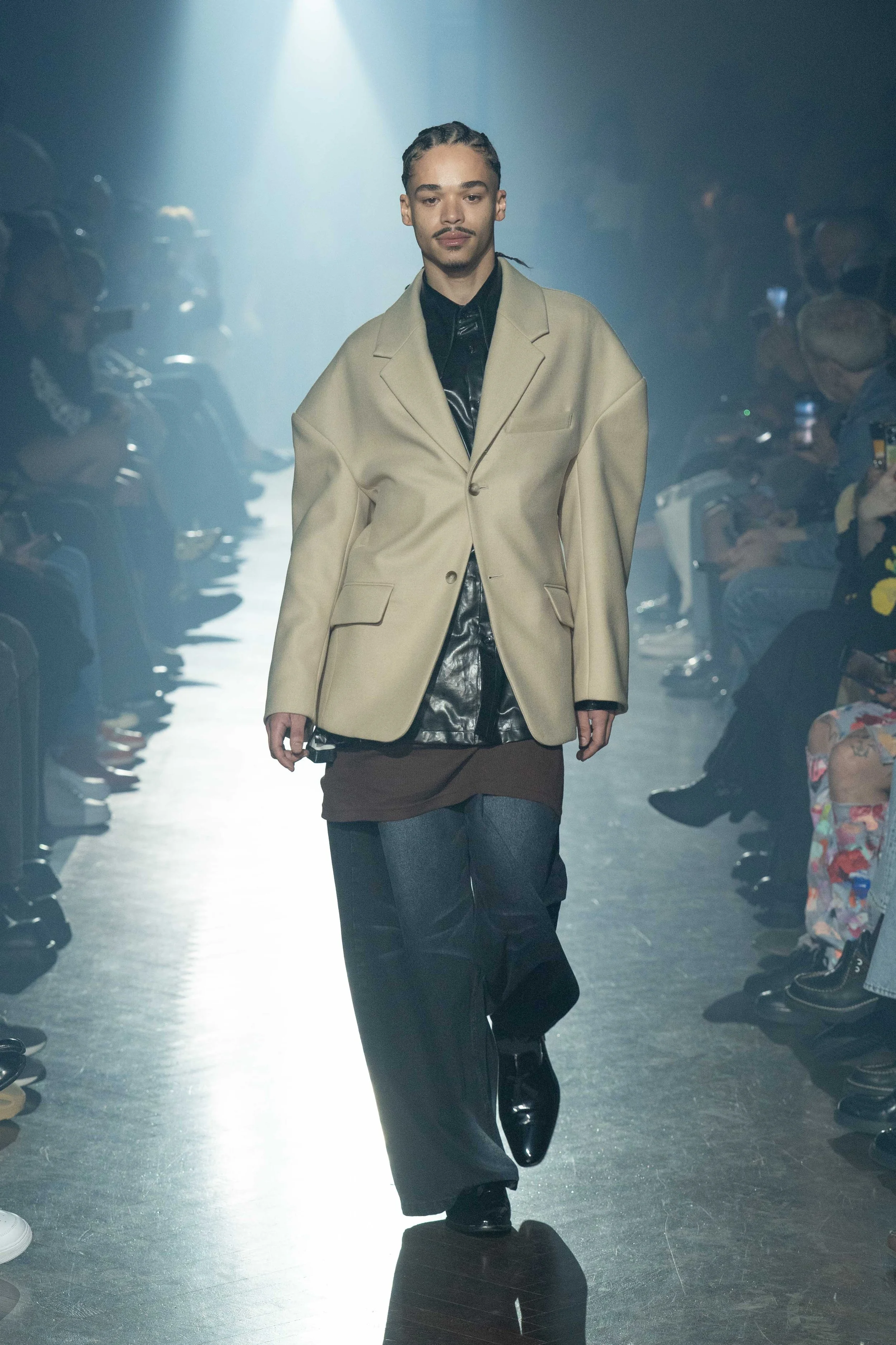 Male model walking on runway during a fashion show wearing a beige oversized blazer, black shiny shirt, brown layered garment, loose dark pants, and black shoes with audience seated on both sides.