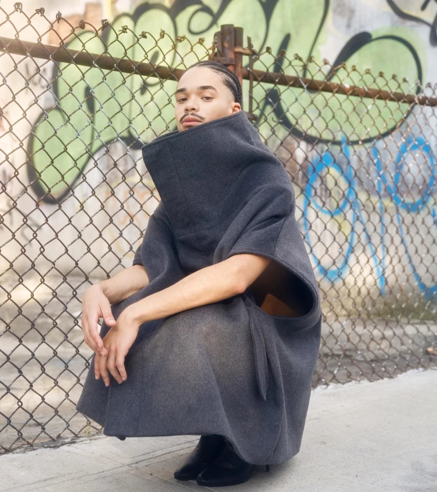 Person squatting in front of a graffiti-covered chain-link fence, wearing a black oversized hoodie with the hood pulled up over their head and black boots.
