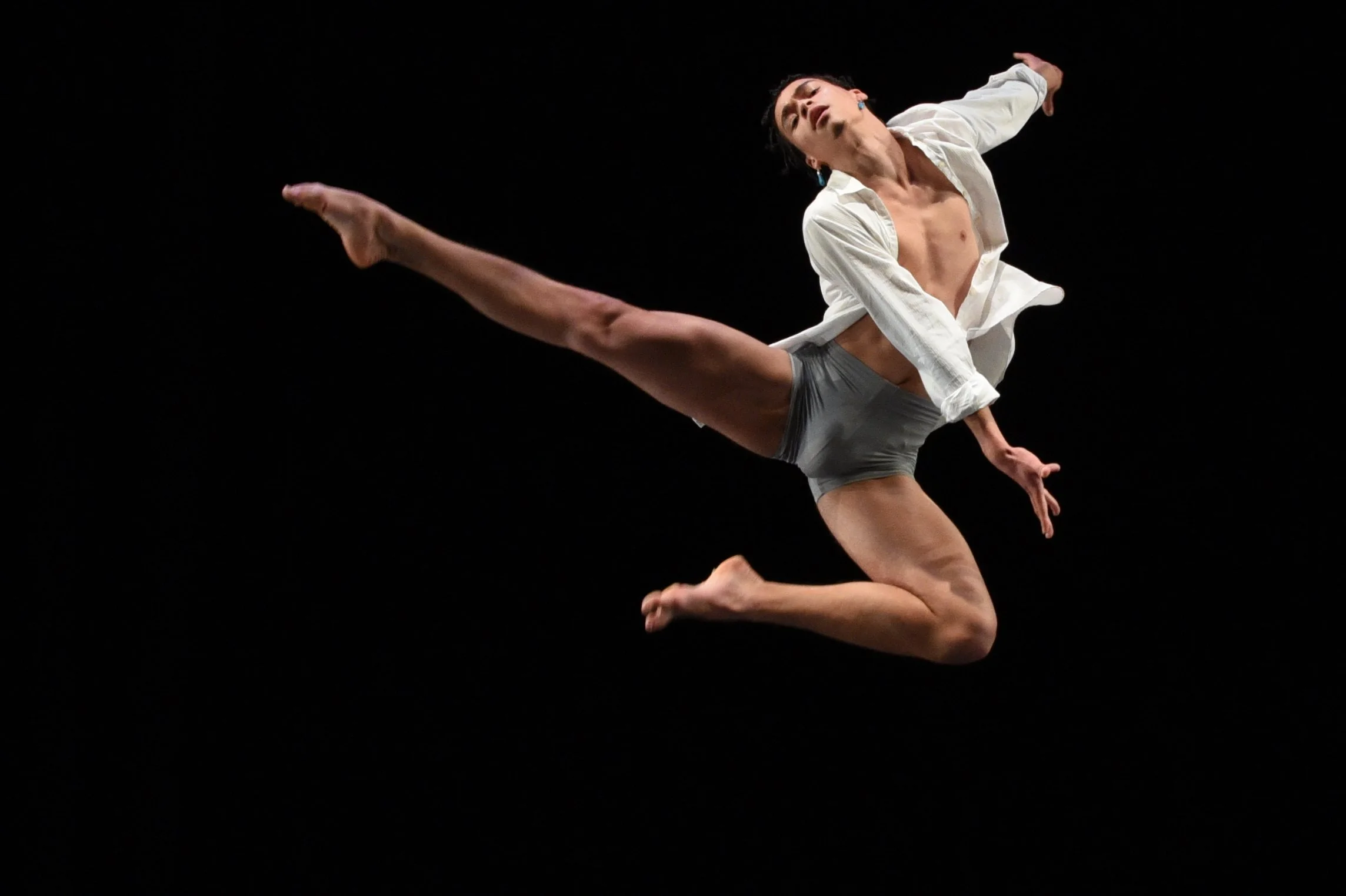 A male dancer in mid-air, performing a leap with one leg extended forward and the other bent backward, wearing a white shirt and gray shorts against a black background.