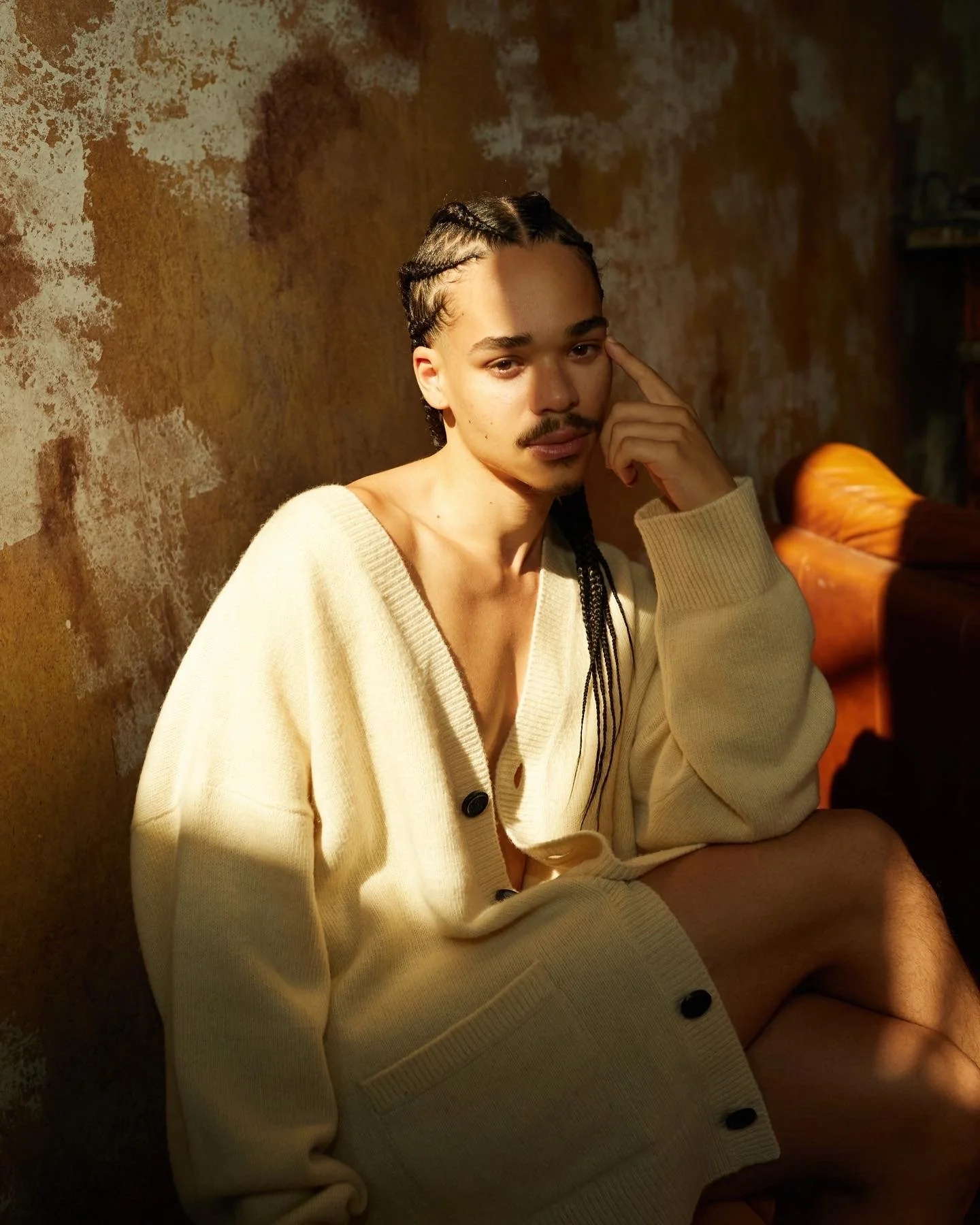 A young man with braided hair, a mustache, and a goatee, sitting on the floor in front of a textured, brown wall. He is wearing a cream-colored, buttoned cardigan that is partly open, revealing his bare chest. He is resting his head on his left hand 
