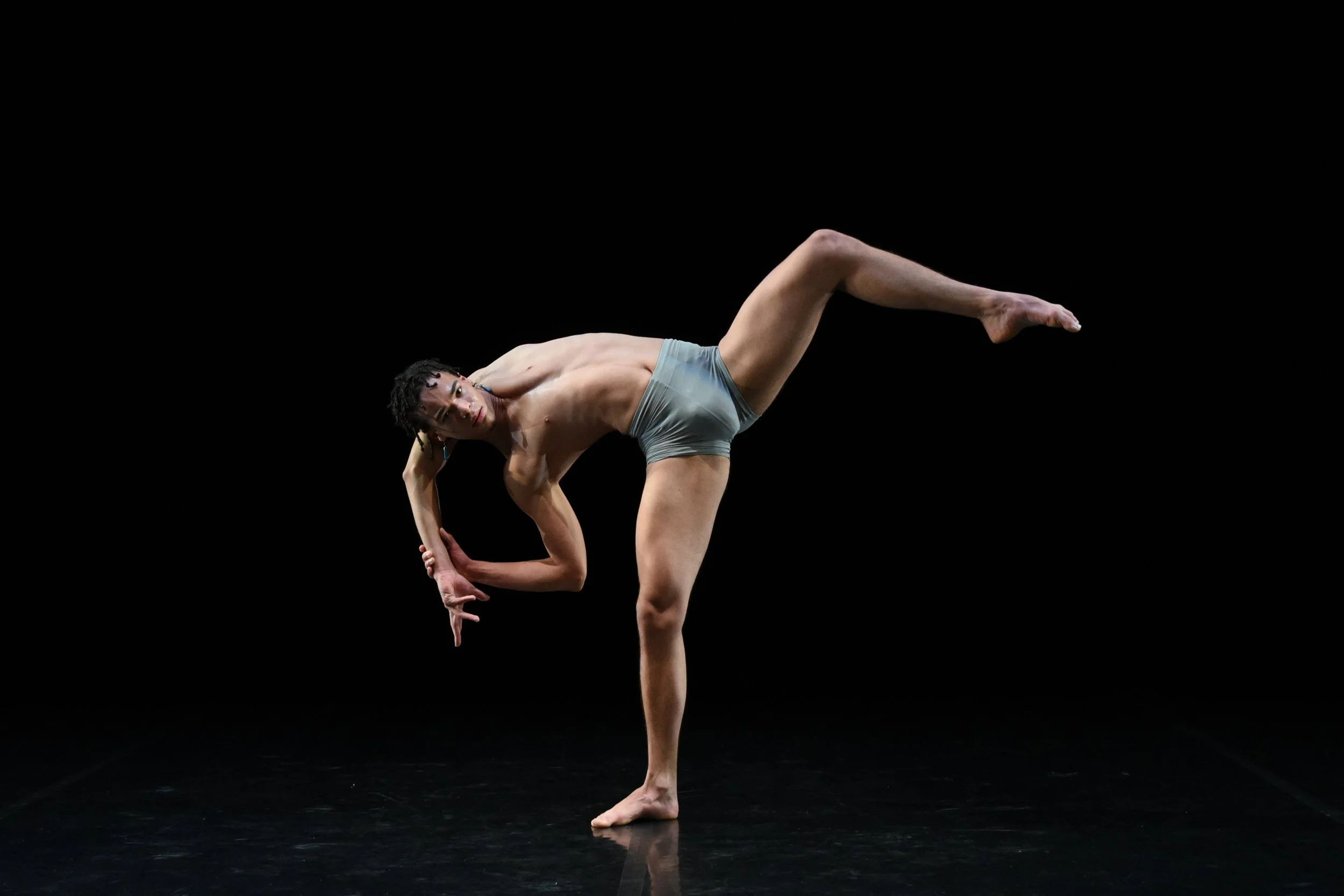 A male dancer performs a pose on stage, balancing on one leg with the other leg extended sideways, and one arm bent behind his head, against a black background.