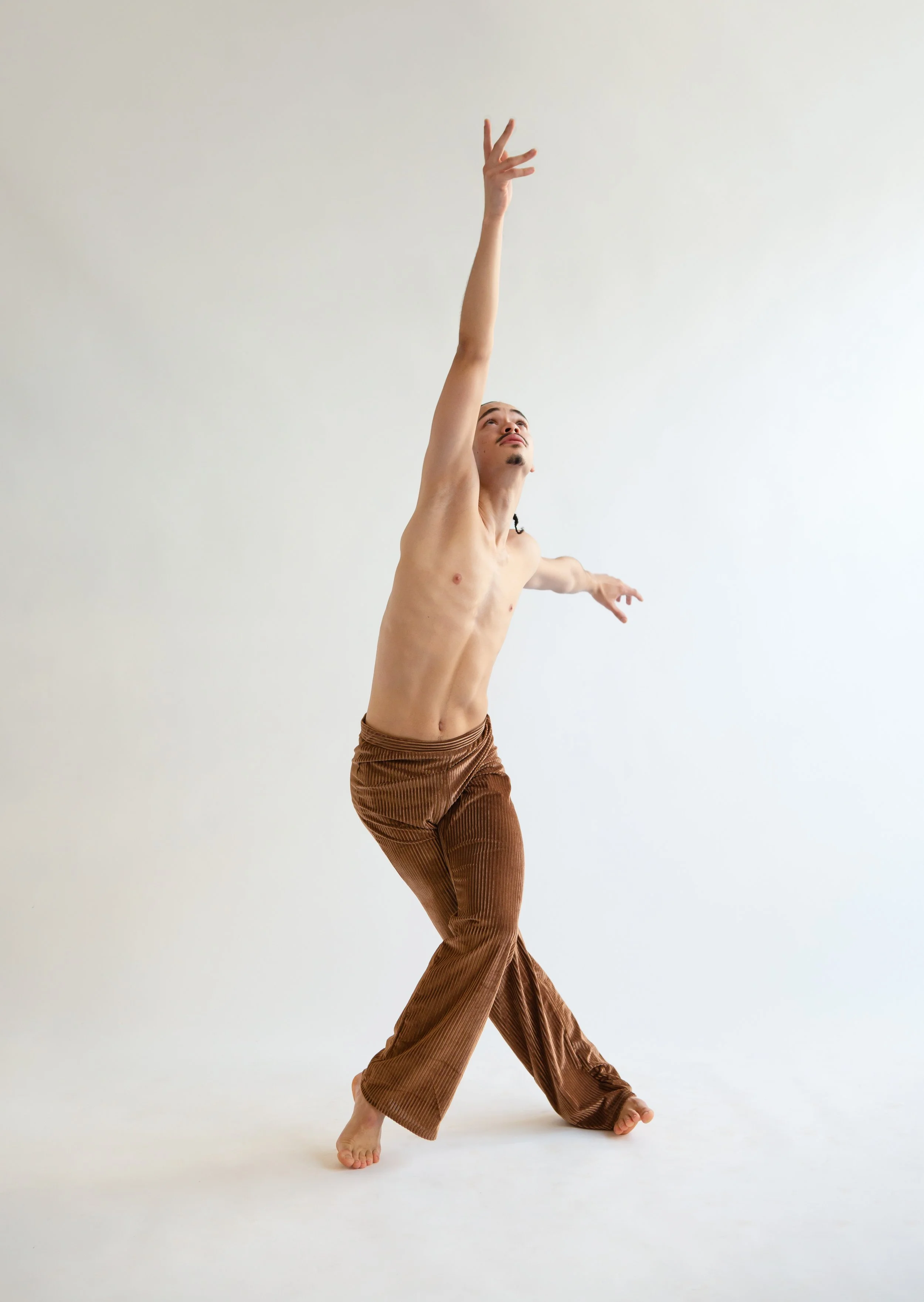 A shirtless man in brown pants poses with one arm extended upward and the other sideways in a dance or dramatic pose on a plain white background.