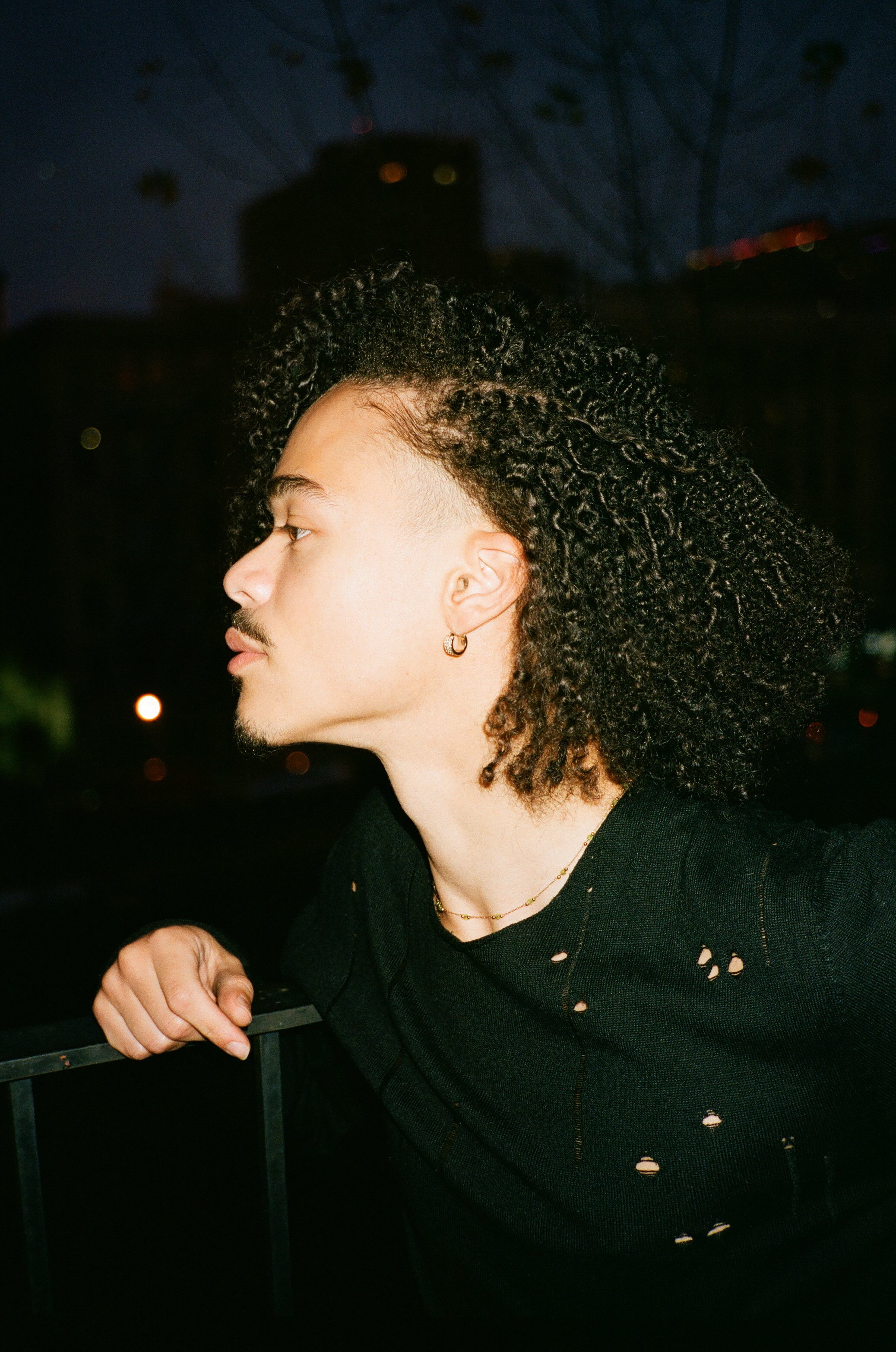 A young man with curly black hair, an earring, and a goatee, standing outdoors at night, looking to the left, wearing a black distressed shirt