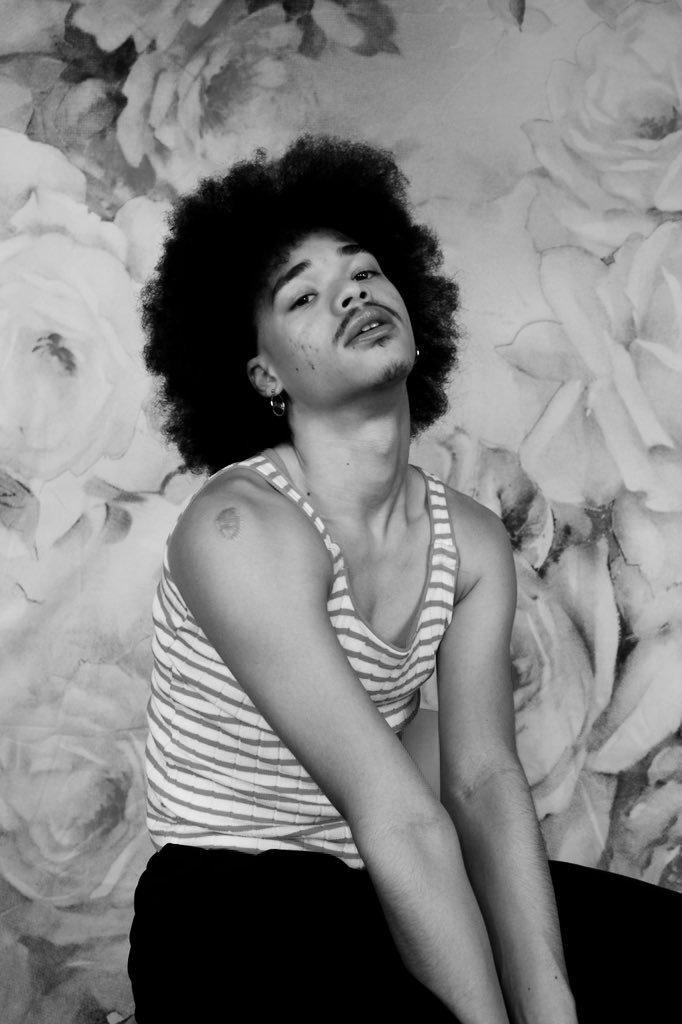 A young man with curly afro hair, wearing a striped tank top, posing with a floral backdrop.
