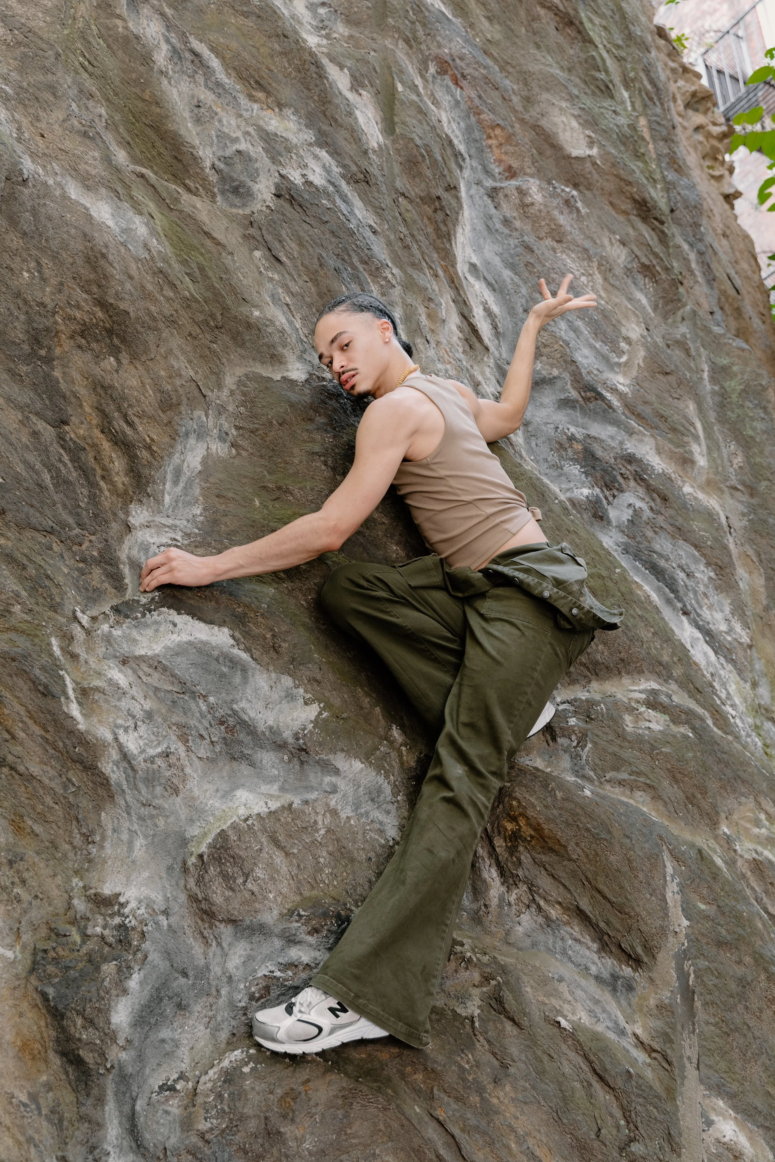 A person with long hair, tank top, and cargo pants is climbing a rugged rock wall outdoors.