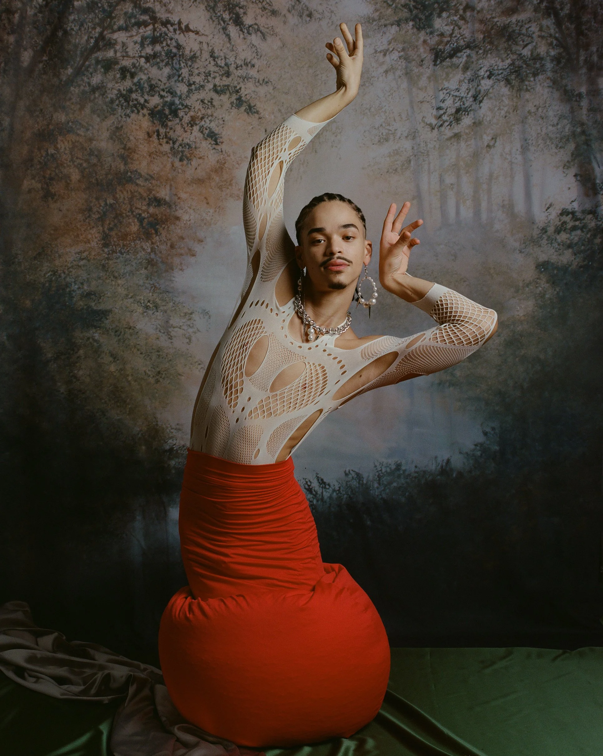 A person with medium skin tone and braided hair, wearing a cream-colored, mesh, cut-out top, a red gathered skirt, and jewelry, poses with arms raised and hands elegantly positioned in front of a painted forest background.