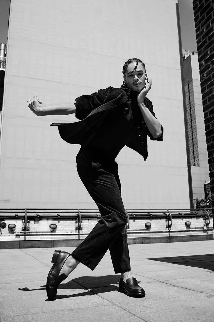 A black and white photo of a woman posing on a city rooftop, dressed in black, with buildings in the background.