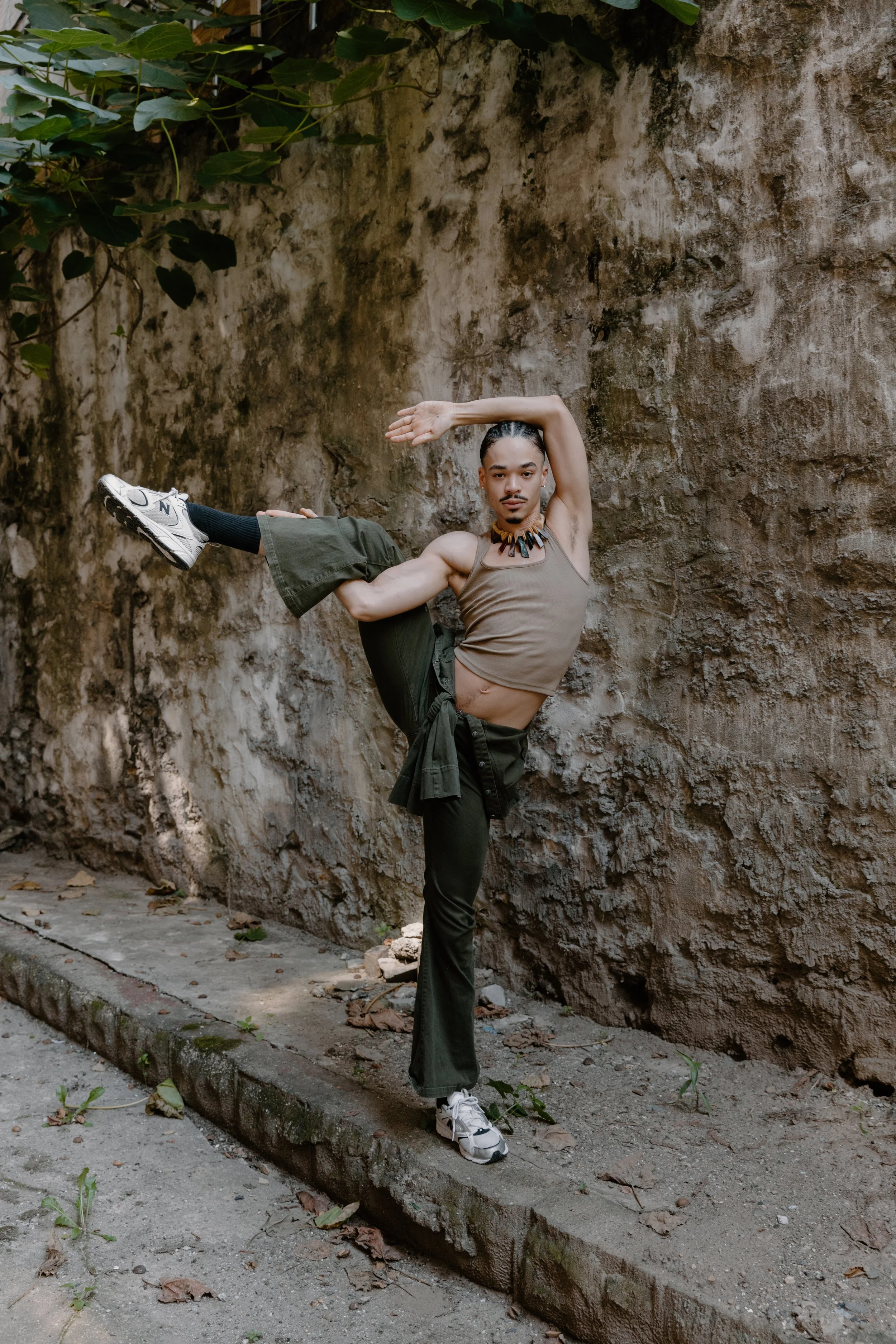 person performing a dance pose against a textured wall, balancing on one leg with a bent knee, and placing one arm over their head and the other across their body, wearing athletic clothing and sneakers.