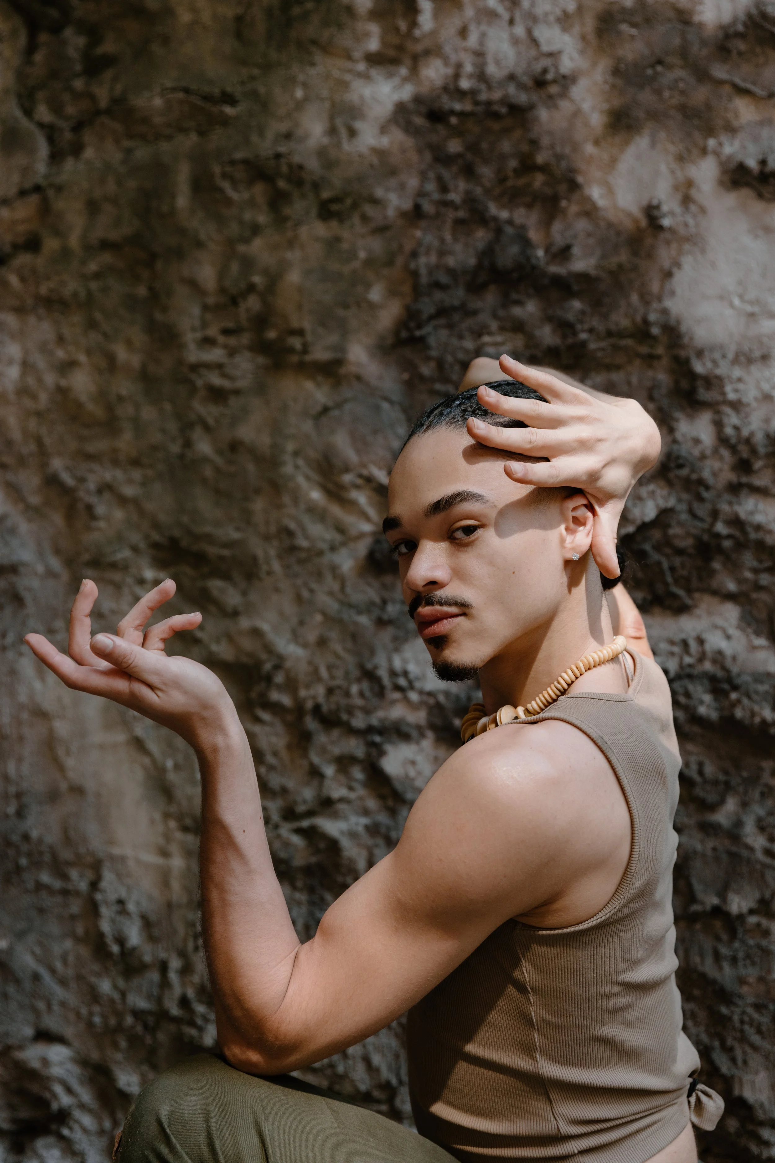 A person with short hair and a goatee, wearing a sleeveless shirt and a shell necklace, posing against a rugged rock wall with one hand near their face and the other raised.