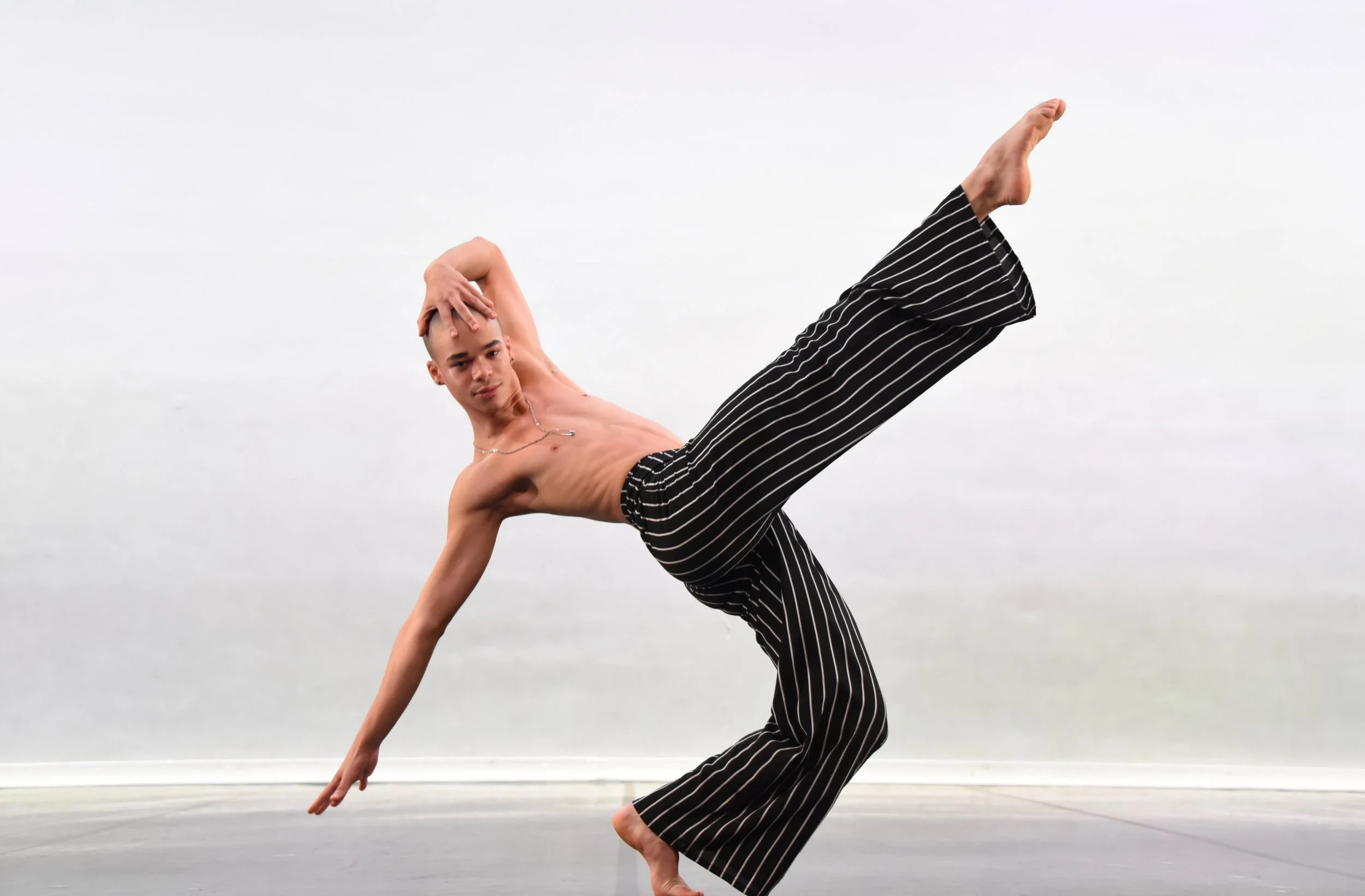 A shirtless young man wearing black and white striped pants is balancing on his right hand with his body sideways, one leg bent and the other extended upwards, on a blank light gray wall background.
