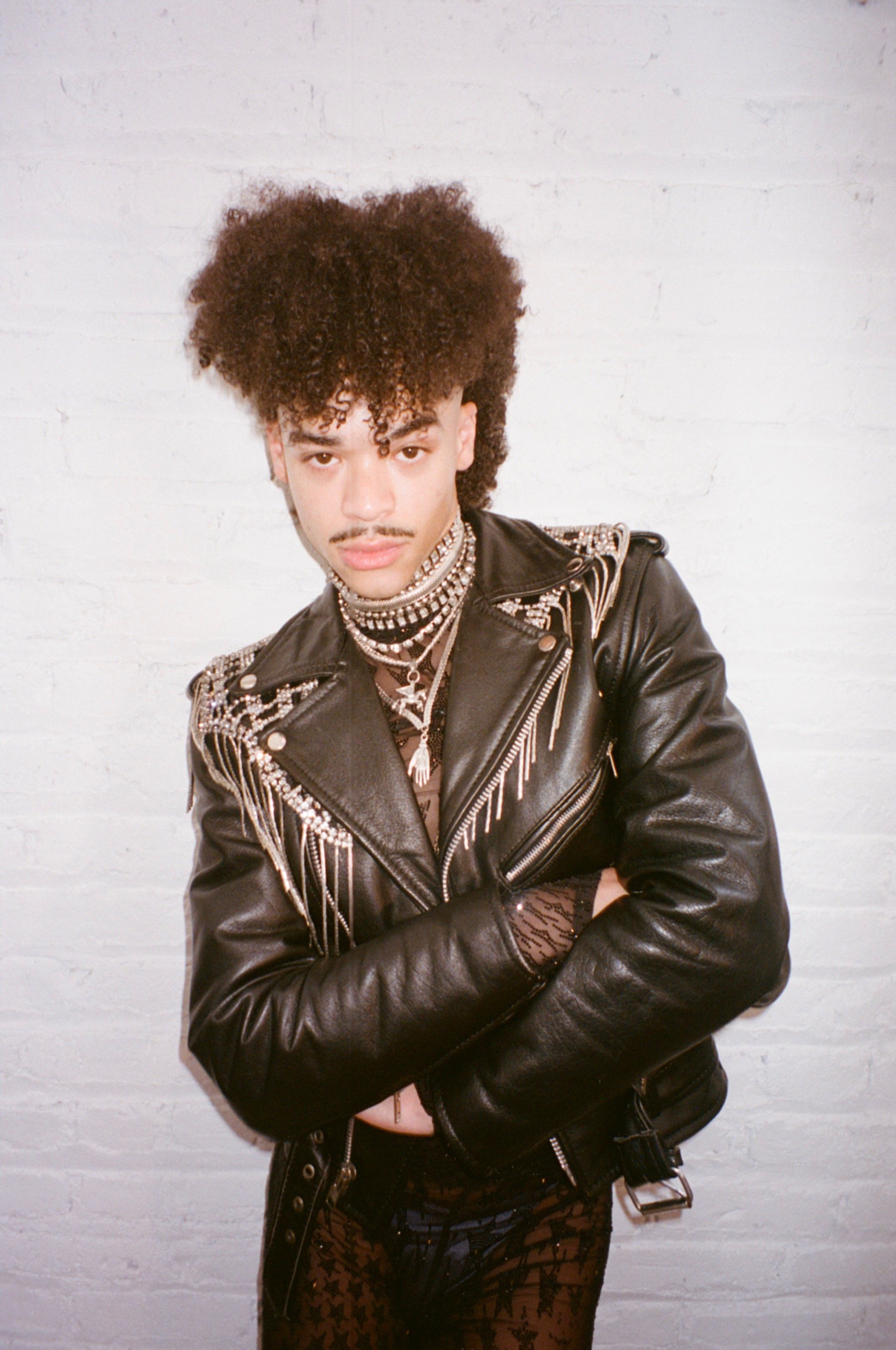 A young person with curly hair and a mustache, wearing a black leather jacket with decorative beads and fringe, layered necklaces, fishnet gloves, and patterned pants, standing against a white brick wall.
