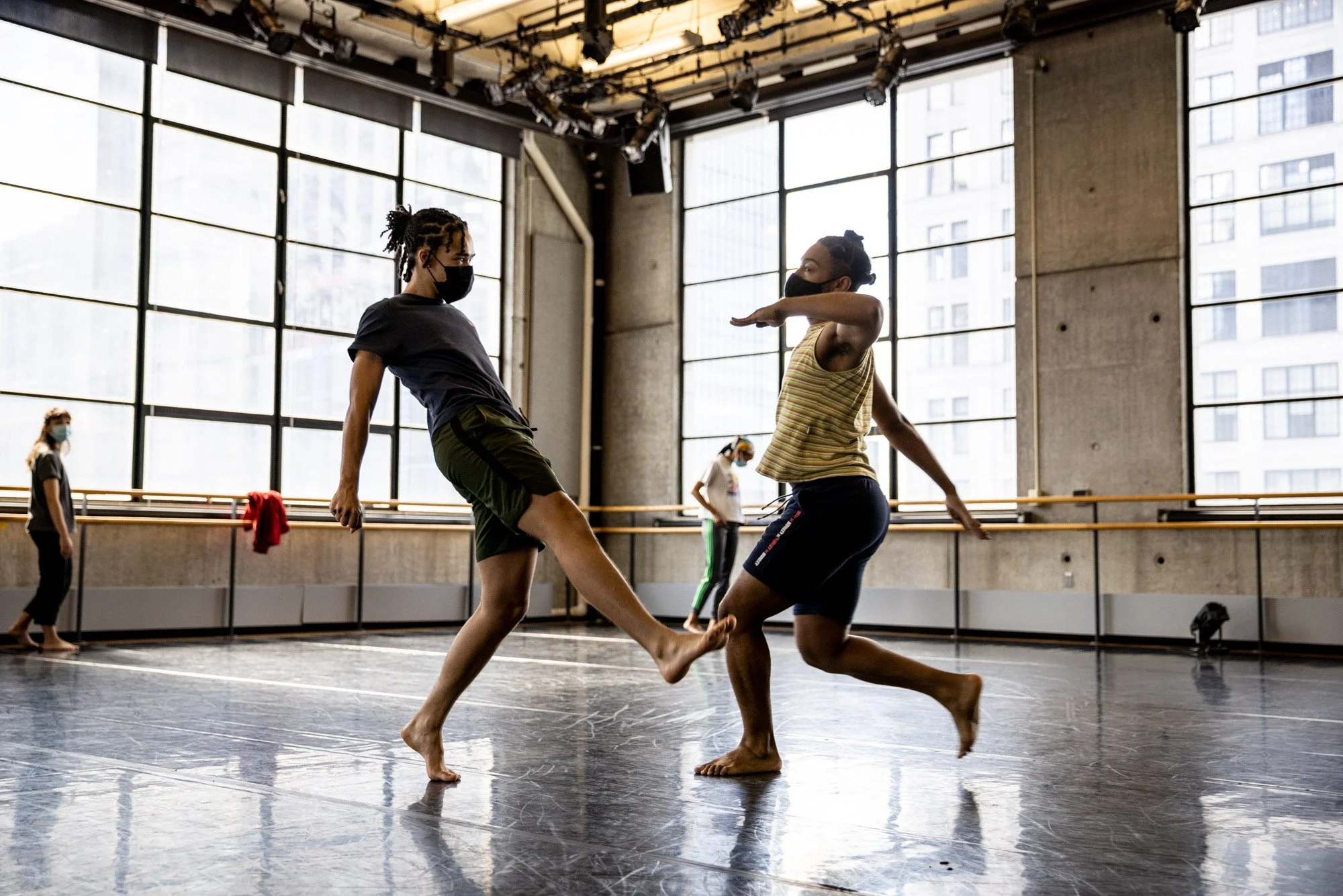 Two dancers wearing face masks practicing a dance routine in a dance studio with large windows, while observing COVID-19 safety measures.