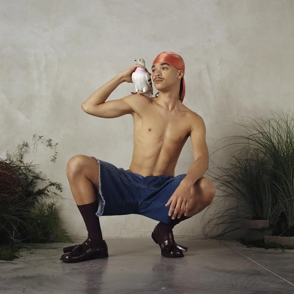 A shirtless young man squatting in front of a plain wall, wearing a pink headscarf, denim shorts, brown socks, and loafers, with a white bird perched on his shoulder and holding another bird near his face amid indoor plants.