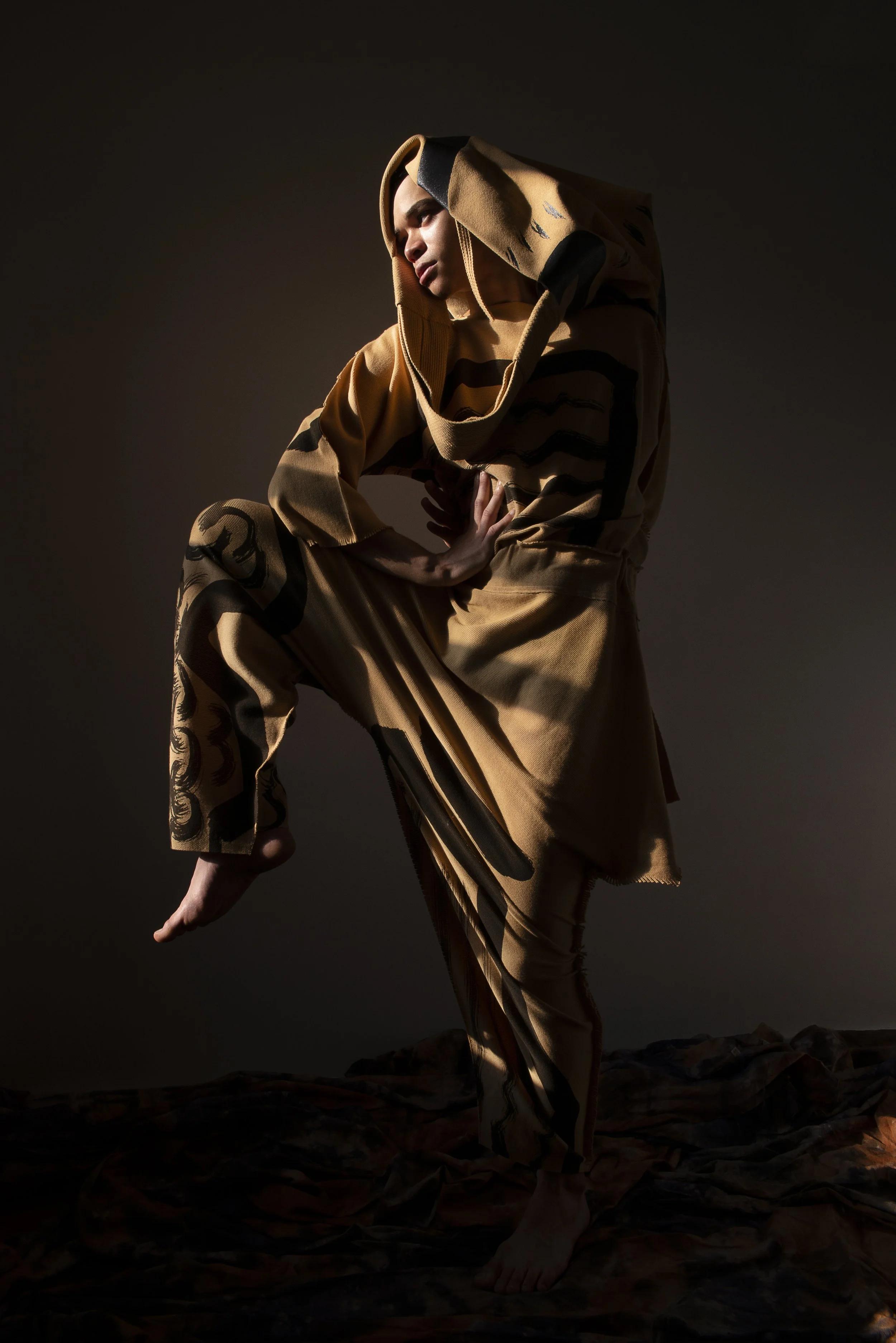 A person dressed in a brown and black patterned hoodie and pants, striking a dramatic pose against a dark background.