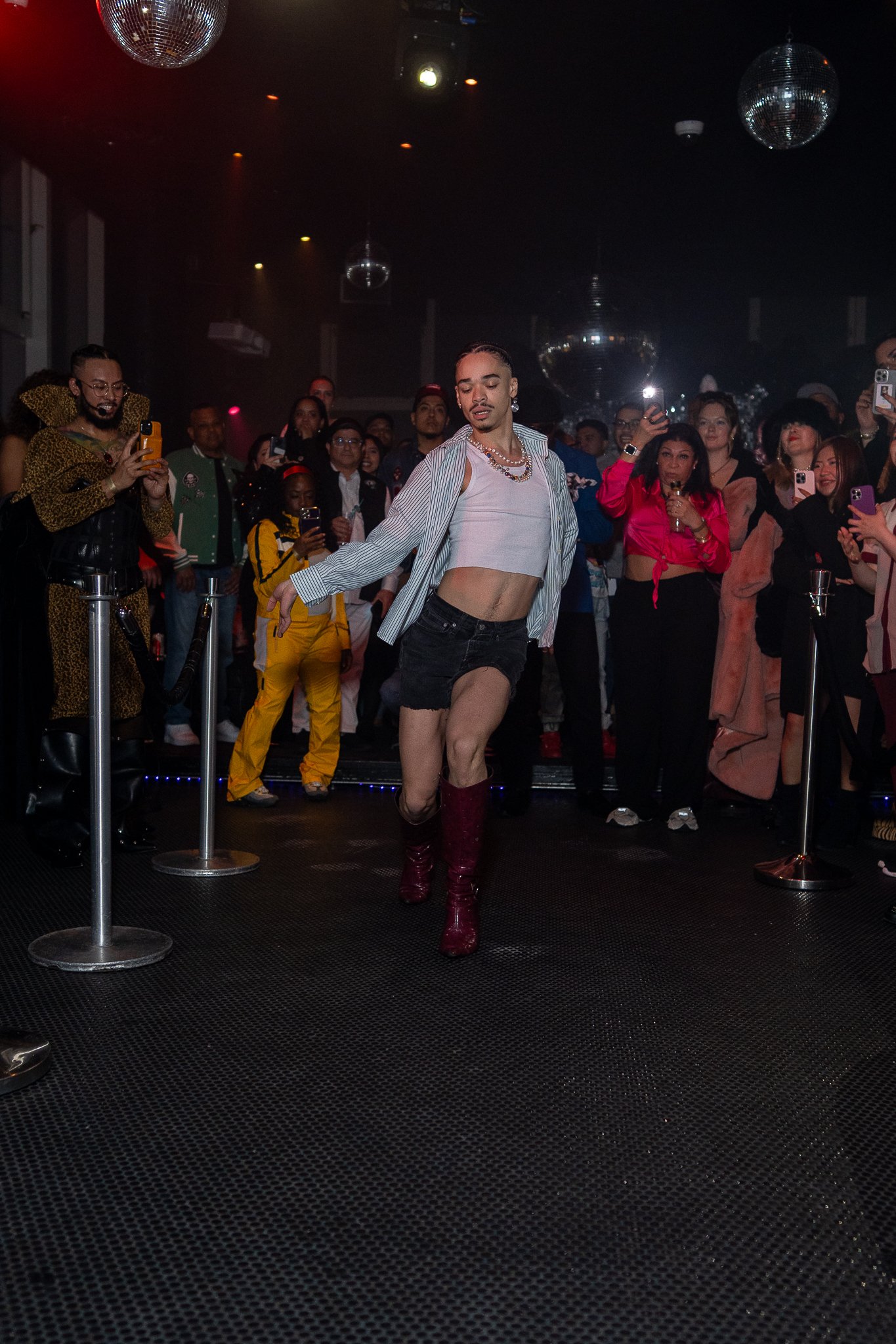 A performer, dressed in a striped shirt, white crop top, black shorts, and red boots, is dancing on a stage with a crowd of onlookers taking photos and videos in the background. The scene appears to be in a nightclub or dance venue with disco balls h
