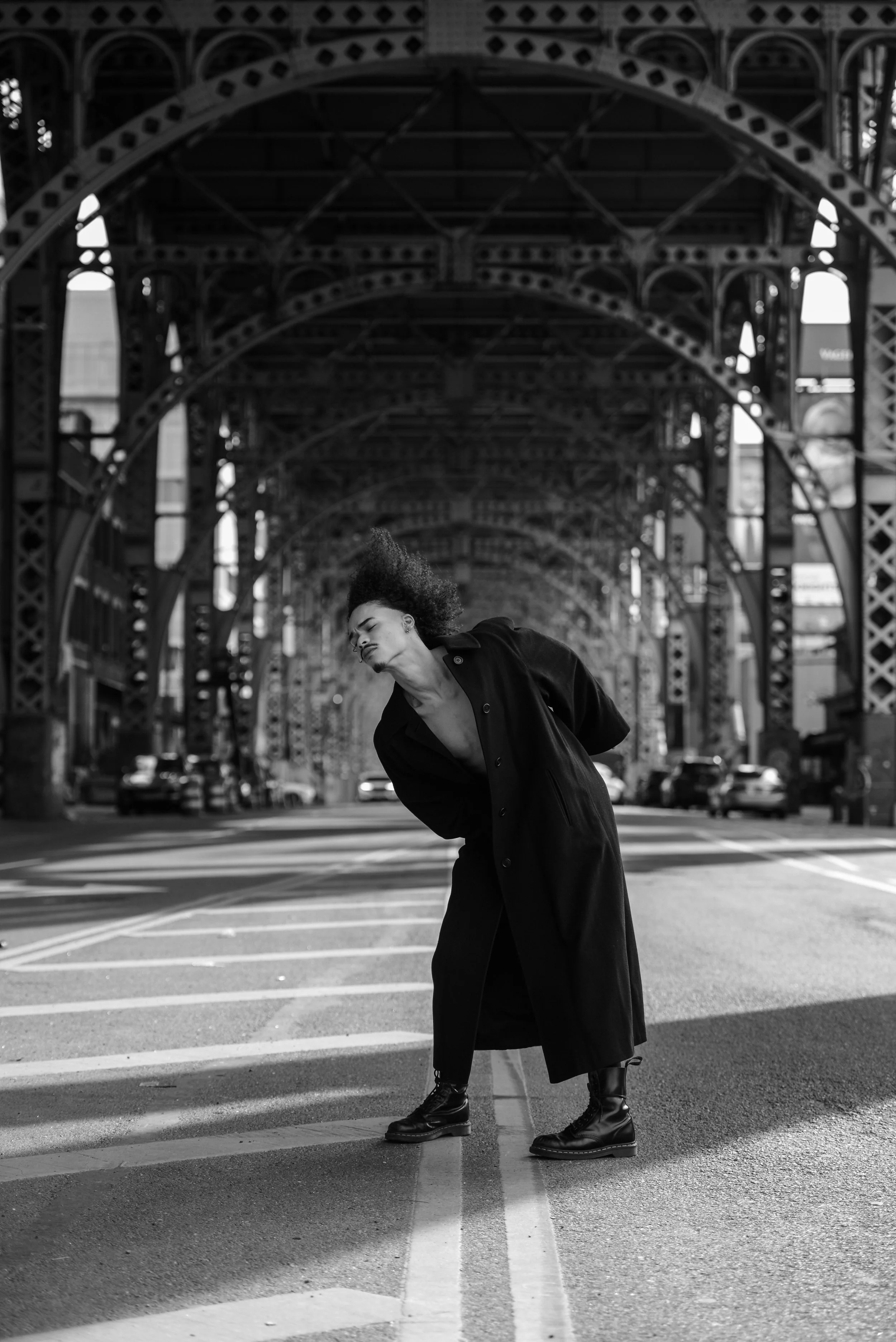 A woman with curly hair wearing a long coat and combat boots, standing in the middle of a city street with an arching bridge overhead, in black and white.