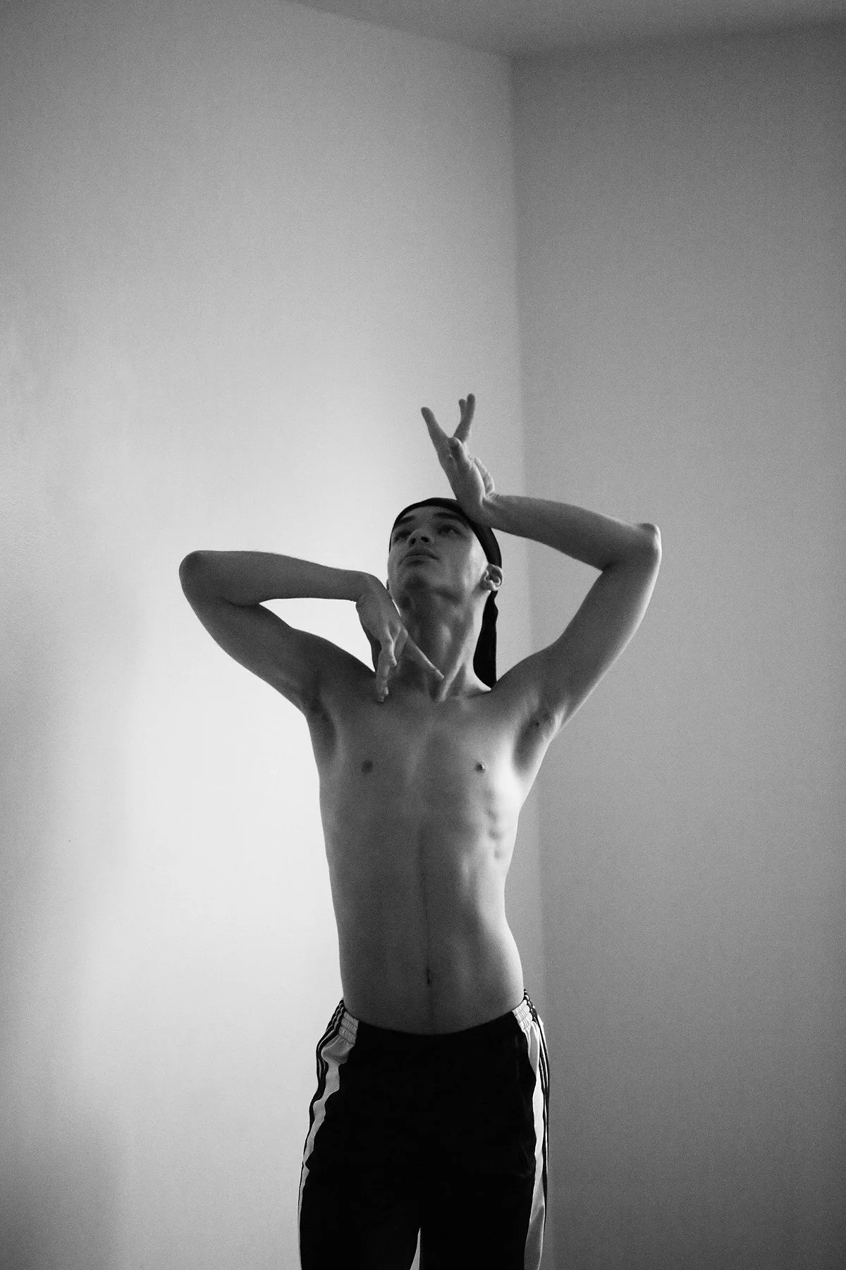 A black and white photo of a young shirtless man with a headband, posing with artistic hand gestures against a plain wall.