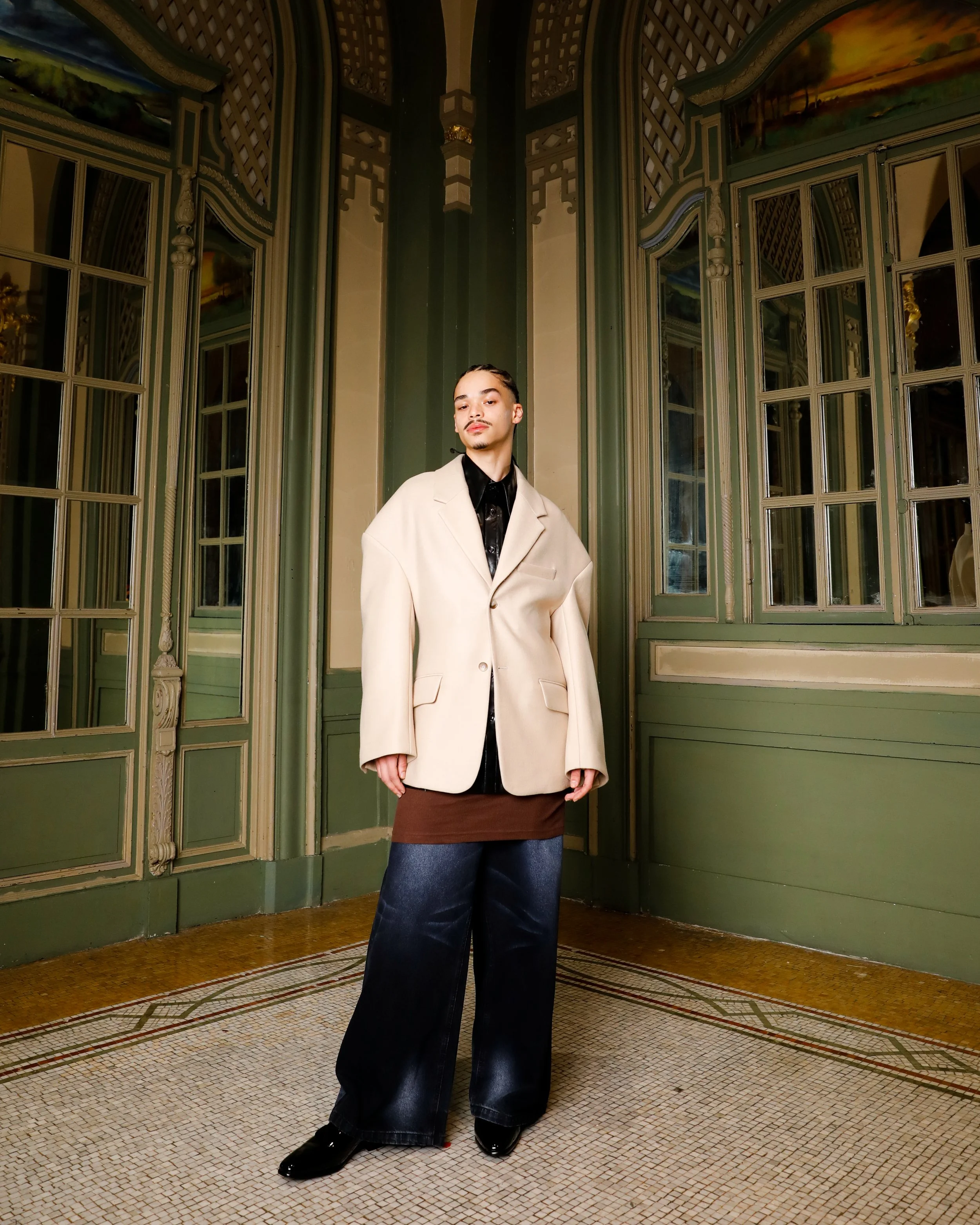 A person standing in an ornately decorated room with large windows and green paneling, wearing an oversized cream blazer, dark shirt, brown skirt, wide-leg jeans, and black shoes.