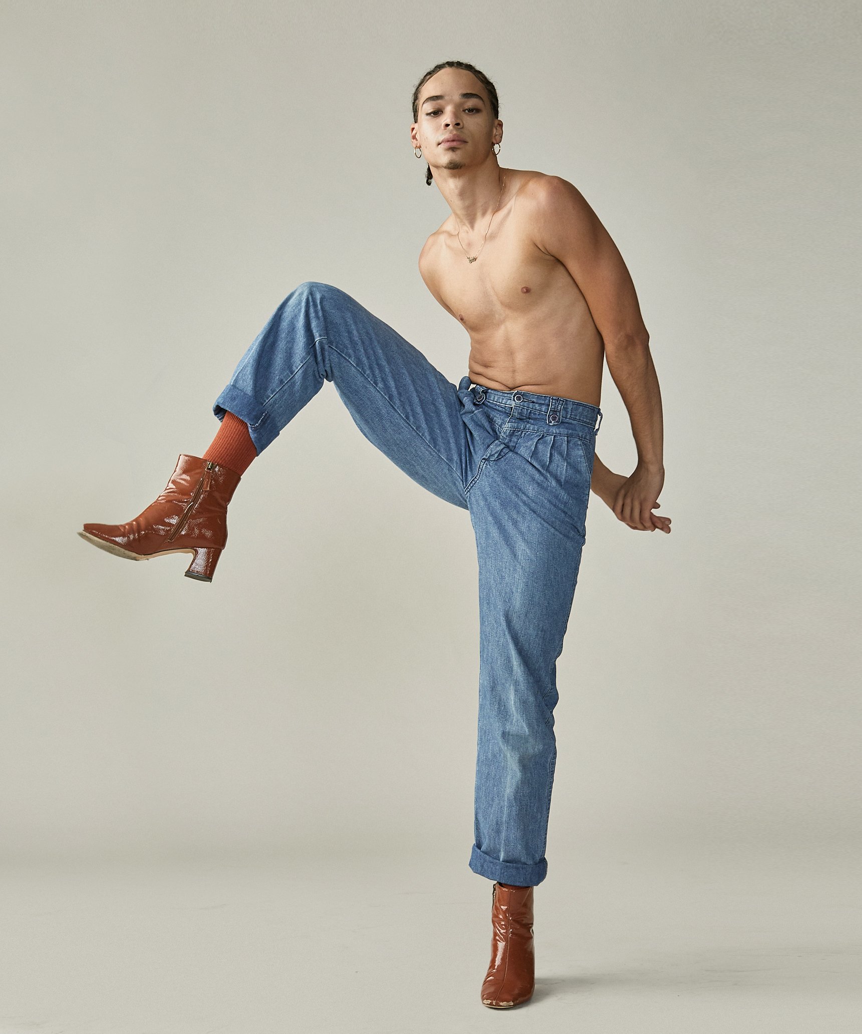 A shirtless young man wearing blue jeans, rust-colored boots, and orange socks, striking a pose against a plain light-colored background.