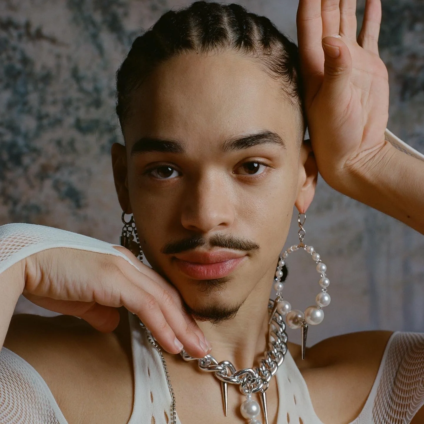 A person with short hair, earrings, and lipstick, posing with their hands near their face against a mottled background.
