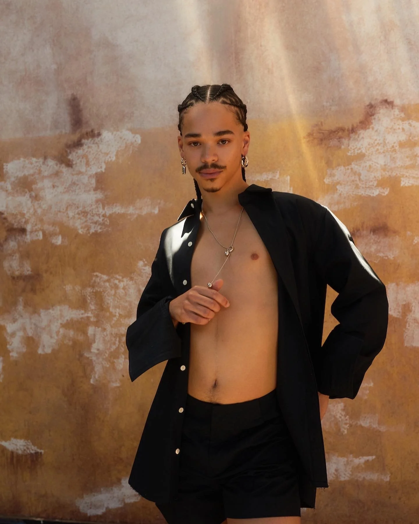A person with braided hair wearing a black shirt open to reveal chest and black shorts, standing against a textured beige background.