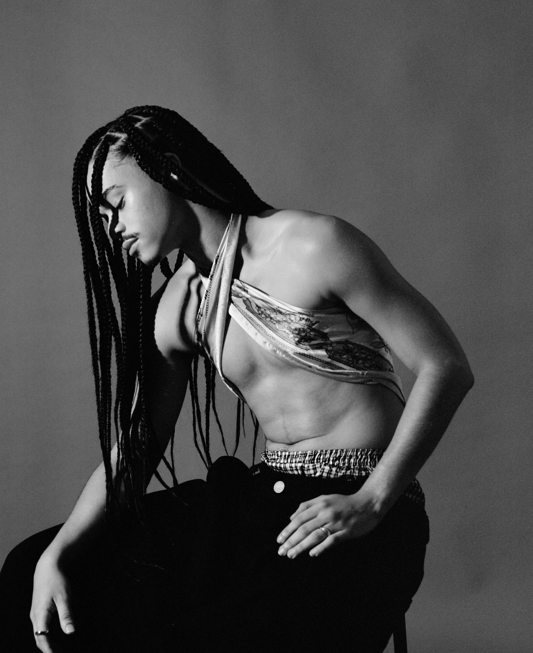 A black-and-white portrait of a woman with long braids, wearing a halter top and sitting with her head bowed and eyes closed.