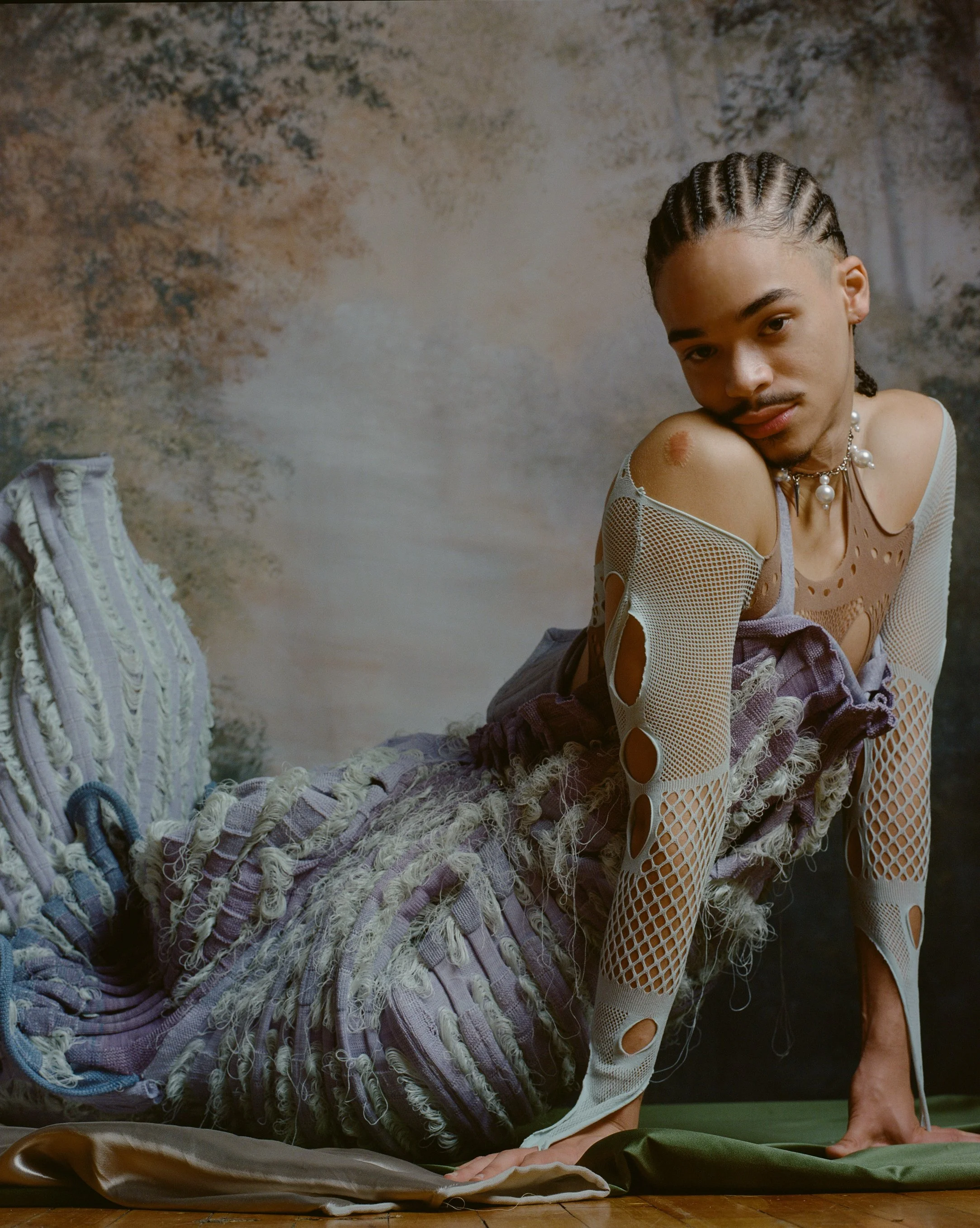 Person with braided hair, wearing mixed fabric clothing with cutouts, is posed on the floor against a textured background.