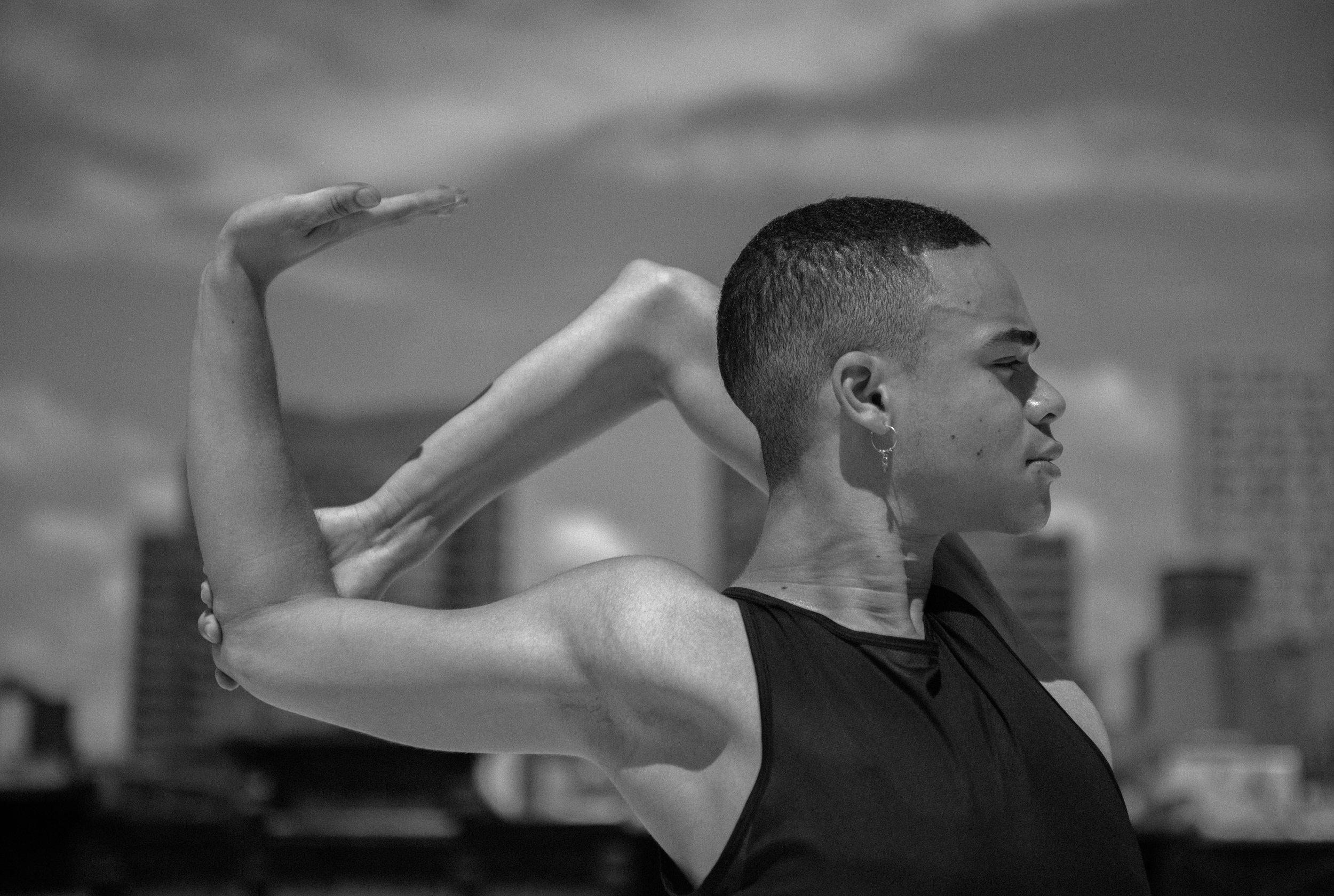 A man with short hair and an earring, wearing a sleeveless top, is outdoor doing a stretch with one arm raised and bent at the elbow while the other arm is touching her back, with city buildings and a cloudy sky in the background.