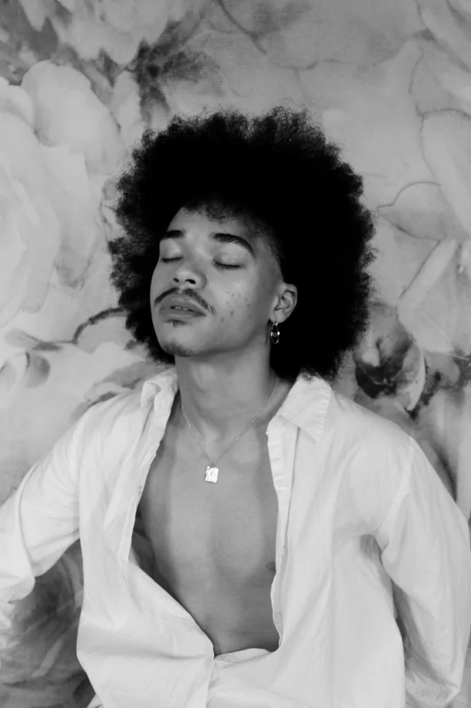 A person with curly afro hair, earrings, and a necklace, wearing an open white shirt, with their eyes closed and a calm expression, standing in front of a floral background in black and white.