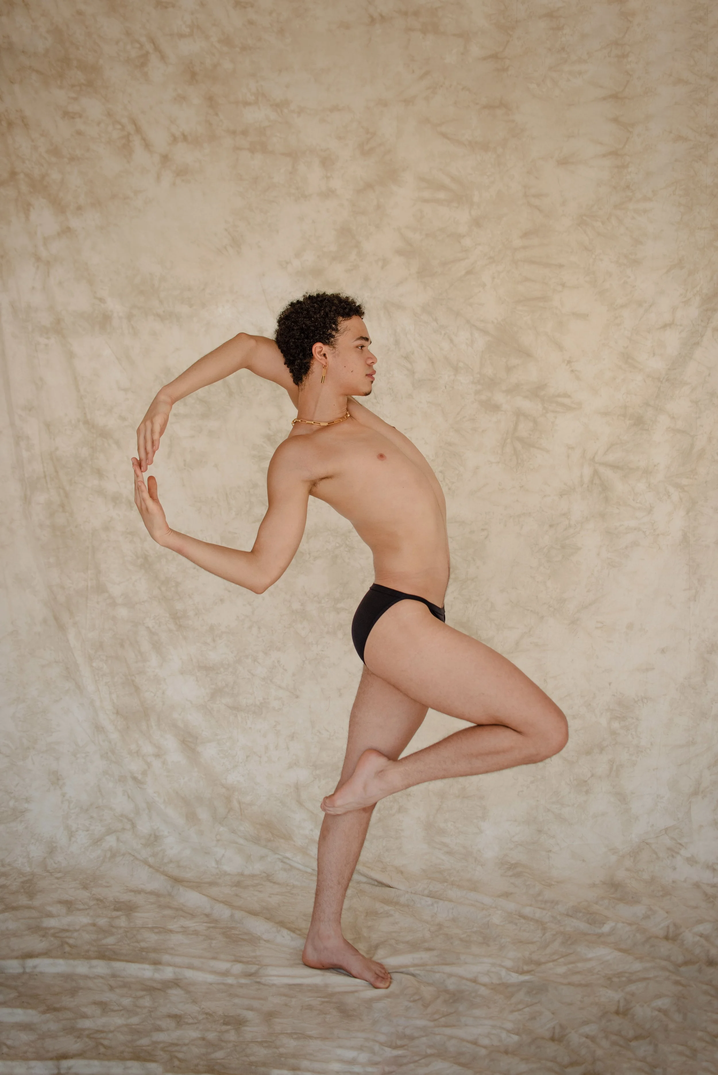 A person with short curly hair and gold jewelry is posing in a dance position against a beige textured background, wearing only black underwear.