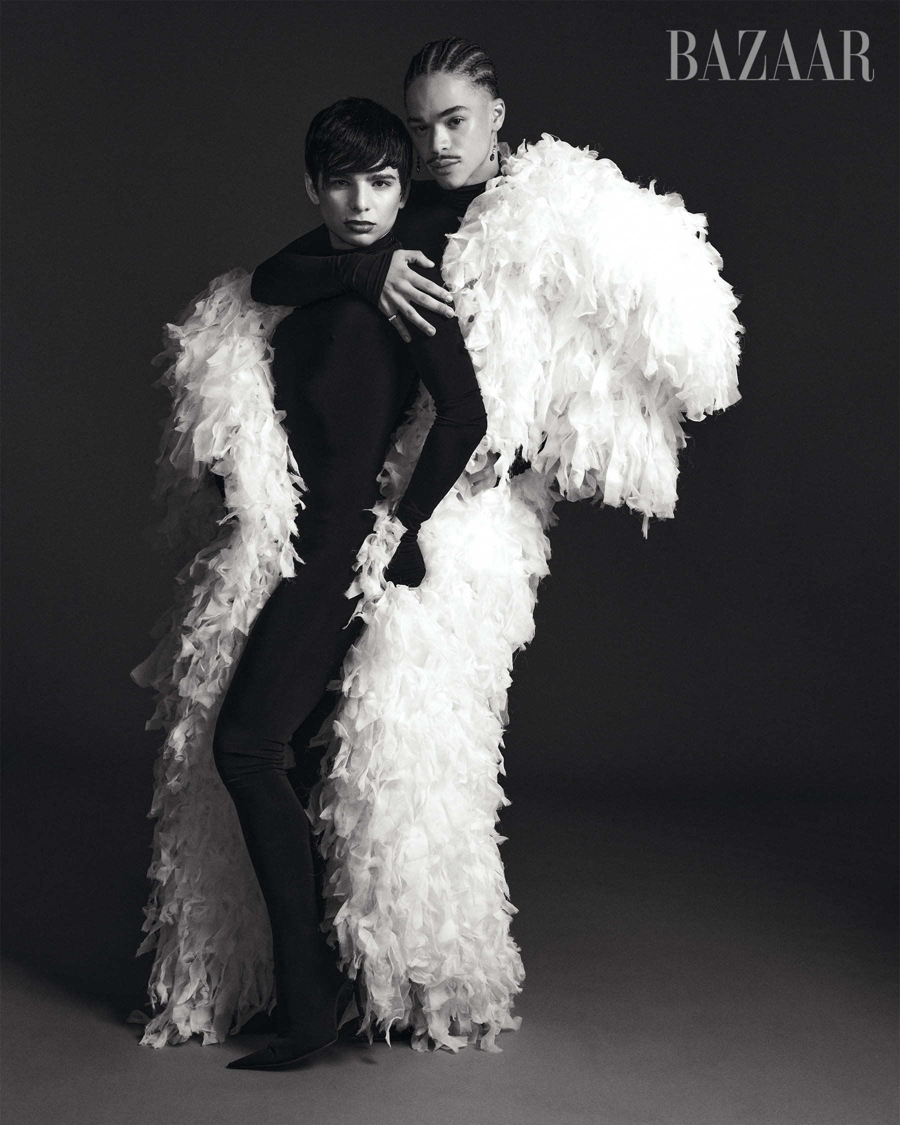 Black and white fashion photo of two models, one with short dark hair and the other with braided hair and facial jewelry, posing together against a plain background. The model with braided hair is wearing a large, feathered white coat, and the other 