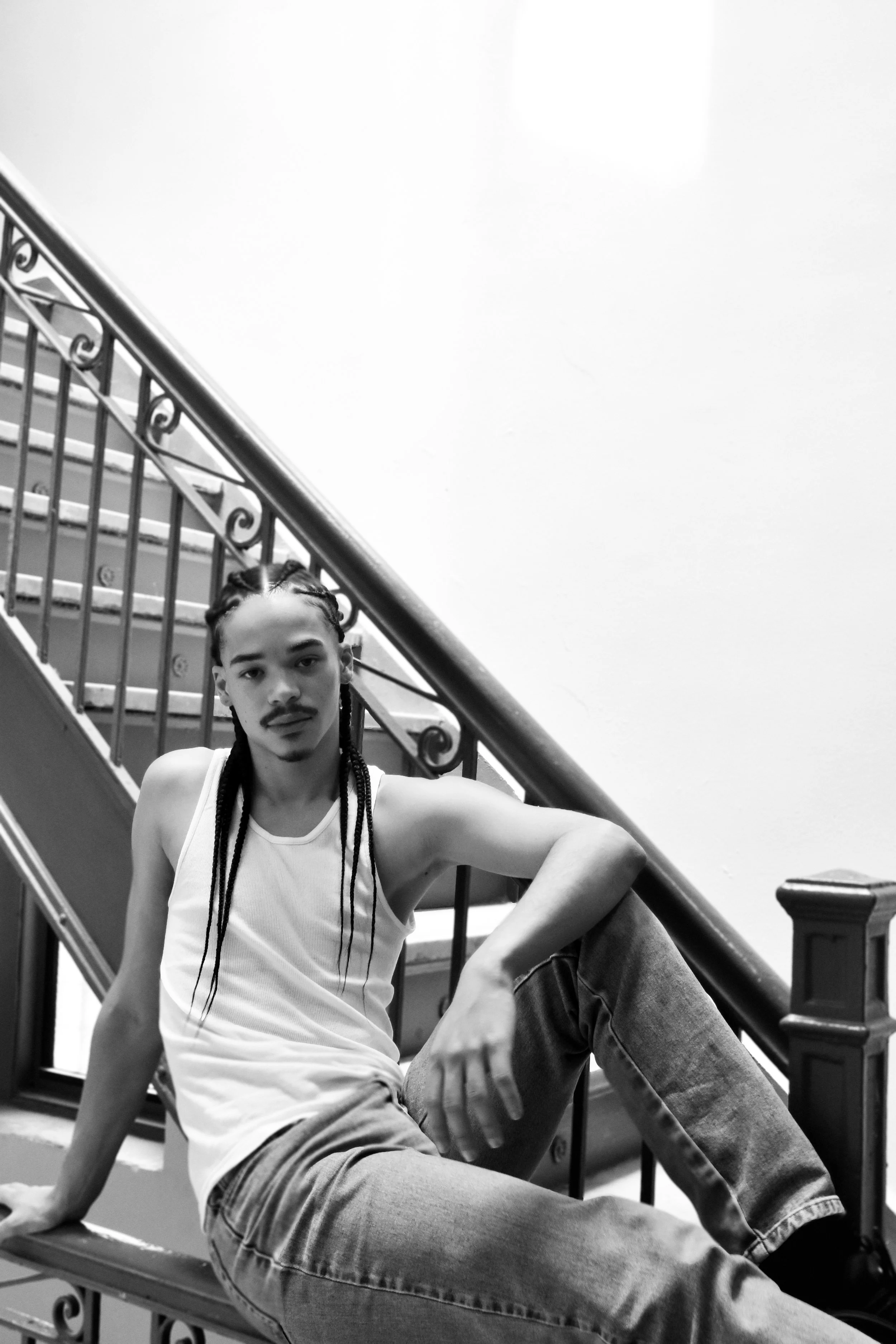 A young man with braided hair and a mustache sitting on a staircase railing, wearing a white tank top and jeans, looking at the camera in black and white.